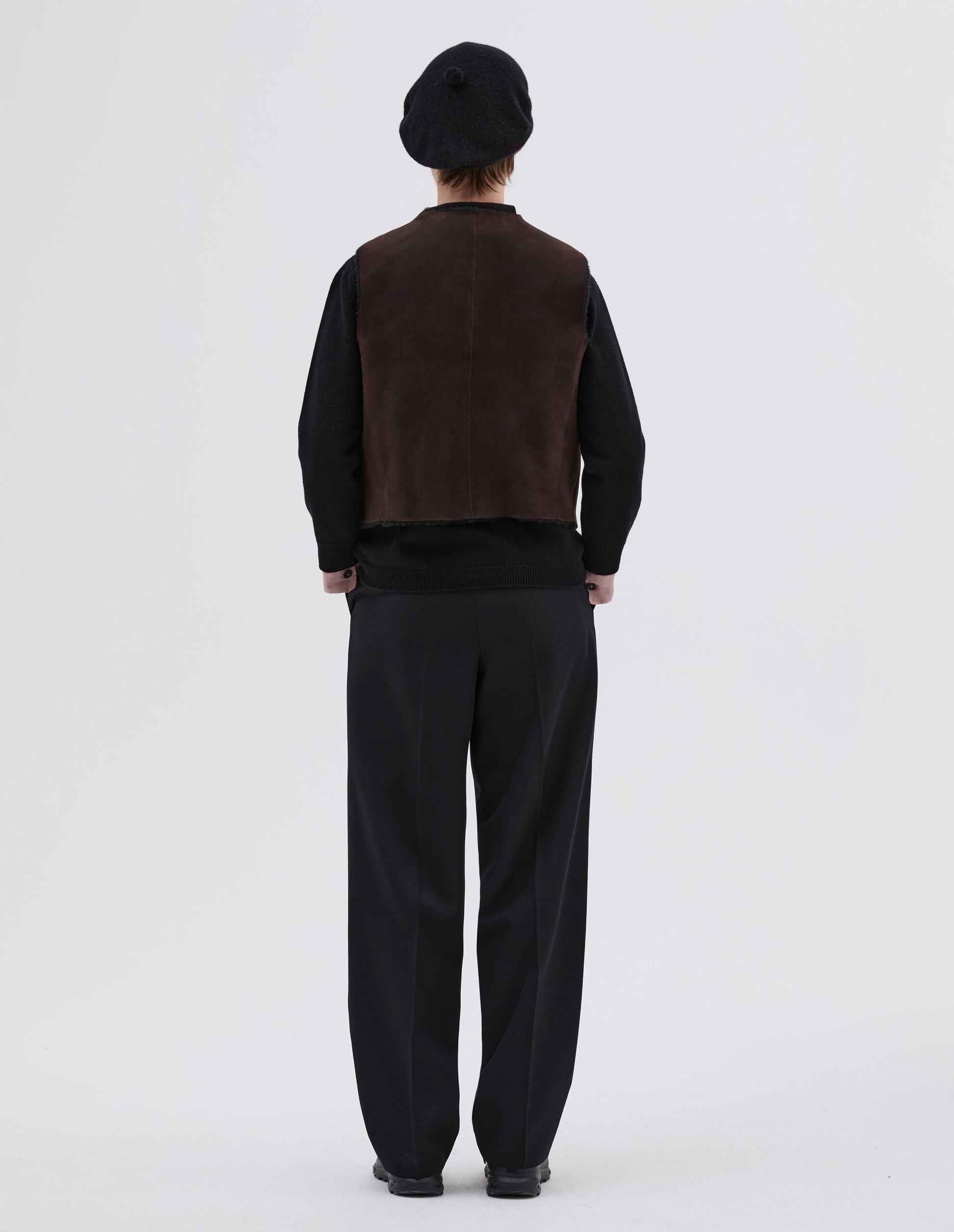 Full body image, back view of model wearing brown and black reversible sleeveless gilet with raw edge finish on neck, armhole and hem. Styled with black beret, black jumper, black trousers and black leather boots.