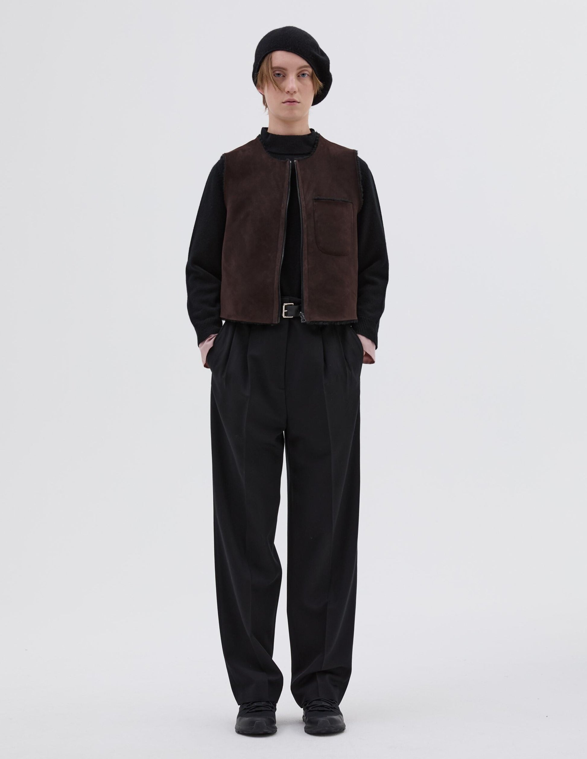 Full body image, front view of model wearing brown and black reversible sleeveless gilet with patch pocket, zip closure and raw edge finish on neck, armhole and hem. Styled with black beret, black jumper, black leather belt, black trousers and black leather boots.