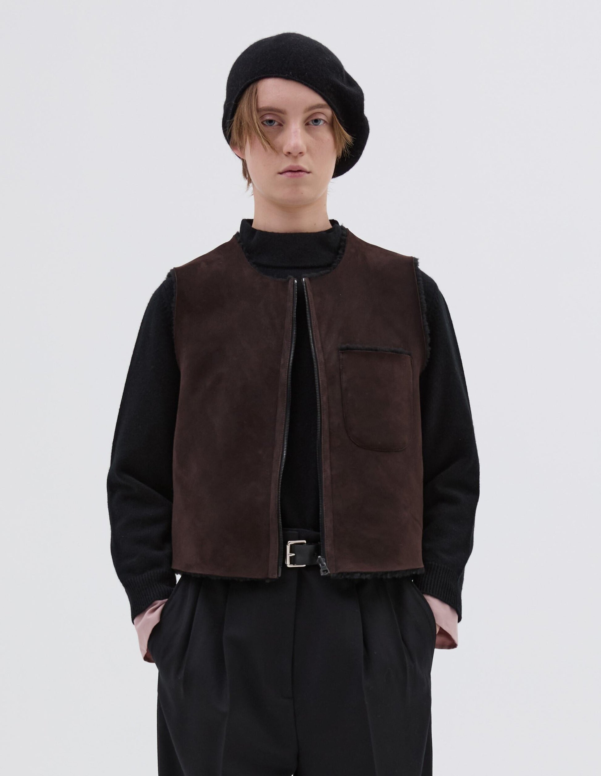 Cropped body image, front view of model wearing brown and black reversible sleeveless gilet with patch pocket, zip closure and raw edge finish on neck, armhole and hem. Styled with black beret, black jumper, black leather belt and black trousers. 