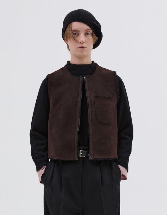 Cropped body image, front view of model wearing brown and black reversible sleeveless gilet with patch pocket, zip closure and raw edge finish on neck, armhole and hem. Styled with black beret, black jumper, black leather belt and black trousers. 