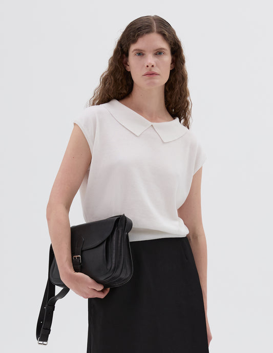 Cropped body image, front view of model wearing black fine grain leather satchel bag with pleat expandable exterior pocket, roller buckle fastening closure and adjustable shoulder strap. Styled with white knitted top and black cotton skirt.