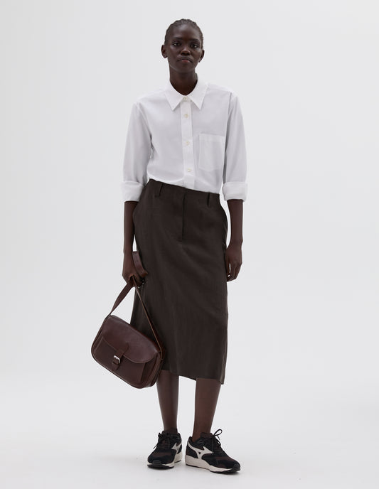 Full body image, front view of model wearing brown fine grain leather satchel bag with pleat expandable exterior pocket, roller buckle fastening closure and adjustable shoulder strap. Styled with white cotton shirt, brown linen skirt and black and brown trainers.