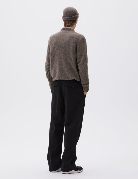 Full body image, back view of model wearing black heavy cotton trousers, with belt loops and concealed and button-through jet back pockets. Styled with brown knitted jumper over a green shirt. With brown knitted beanie hat and black and brown trainers.