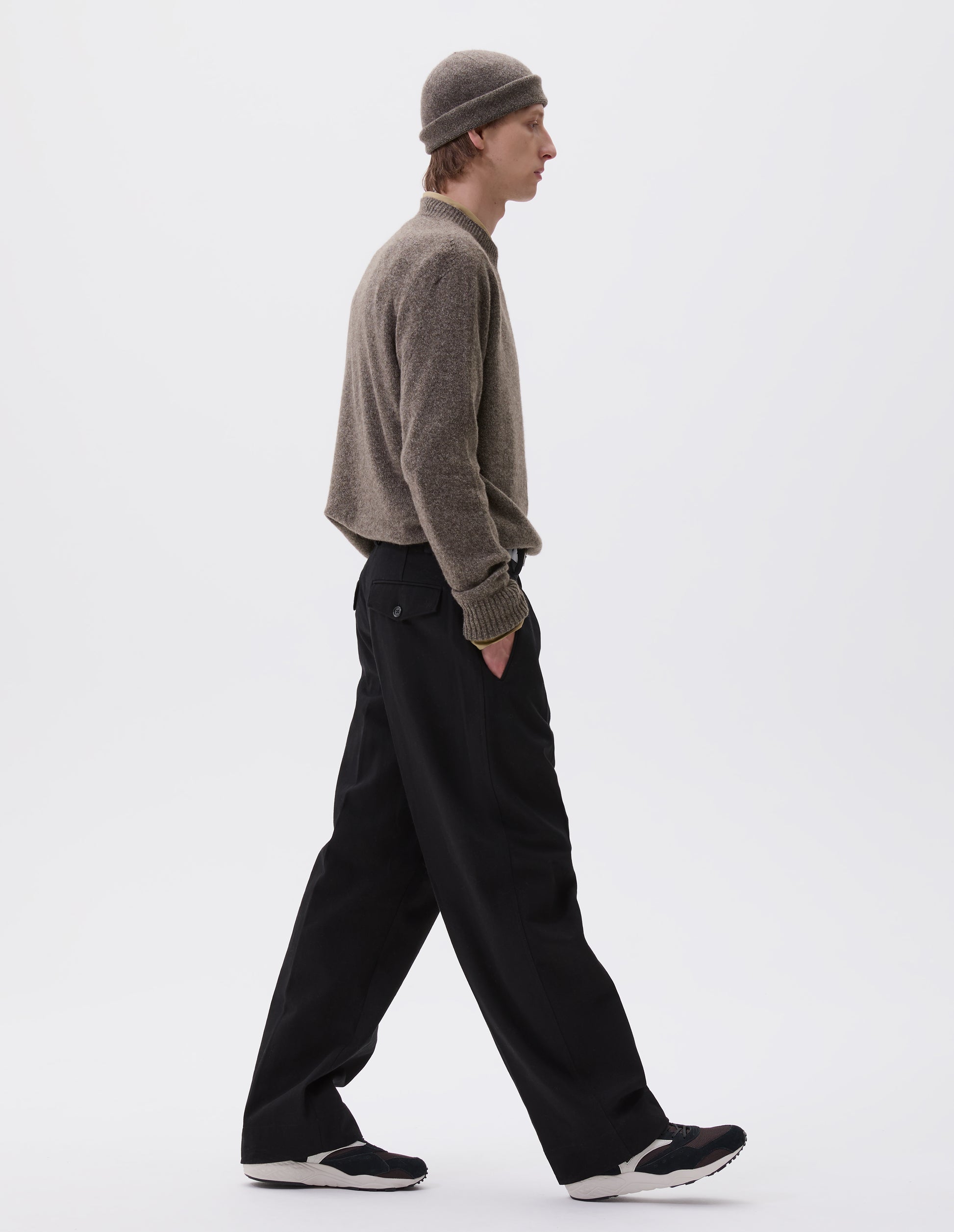 Full body image, side view of male model wearing a pair of black and dark brown suede trainers with mesh insert and off white sole, with off white Mizuno logo running through the side. Styled with beige jumper and matching hat and black trousers.