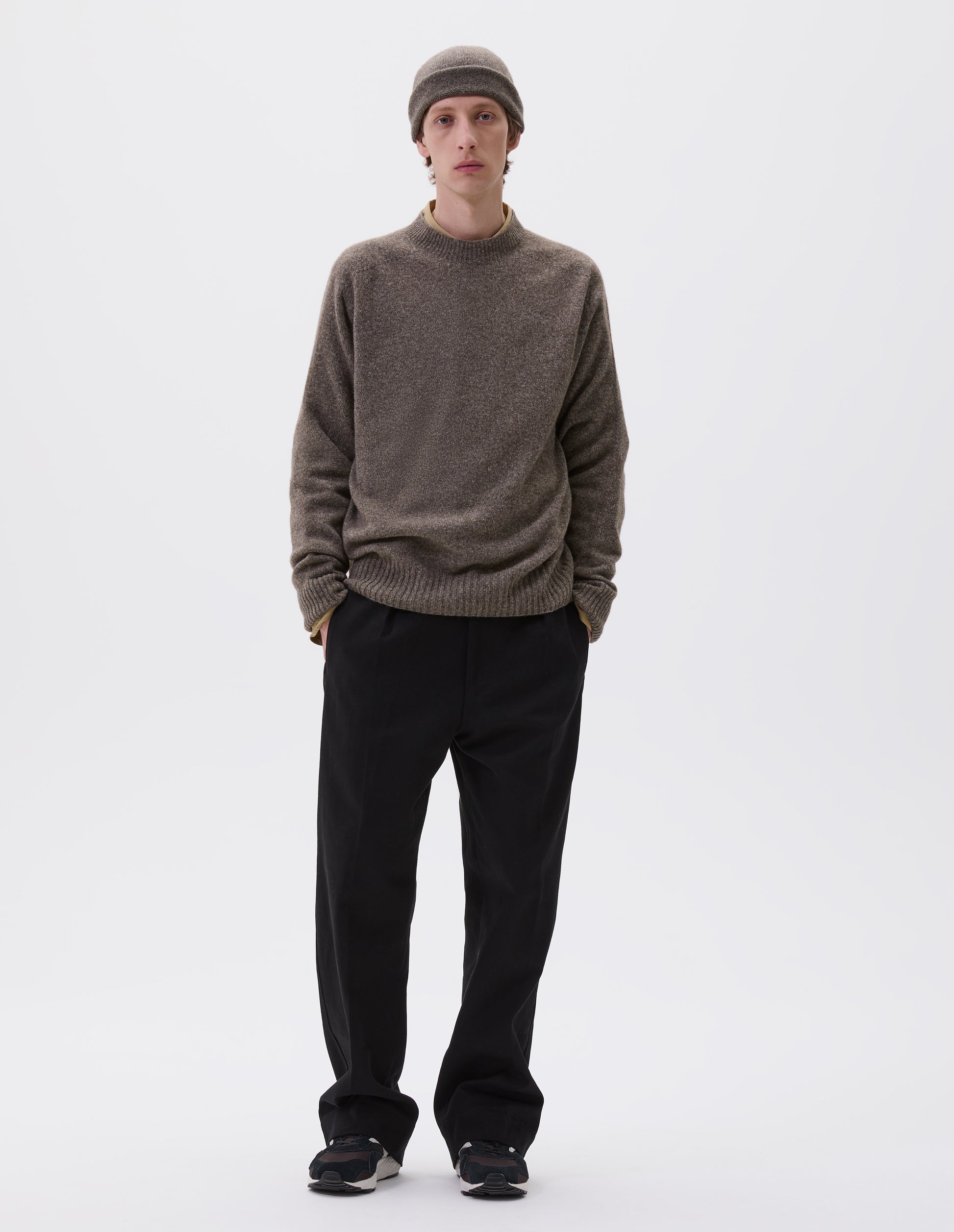 Full body image, front view of model wearing beige marl long sleeve jumper with contrast rib neckline, cuffs and hem. Styled over a yellow shirt, black trousers and with matching knitted hat and black and brown trainers.