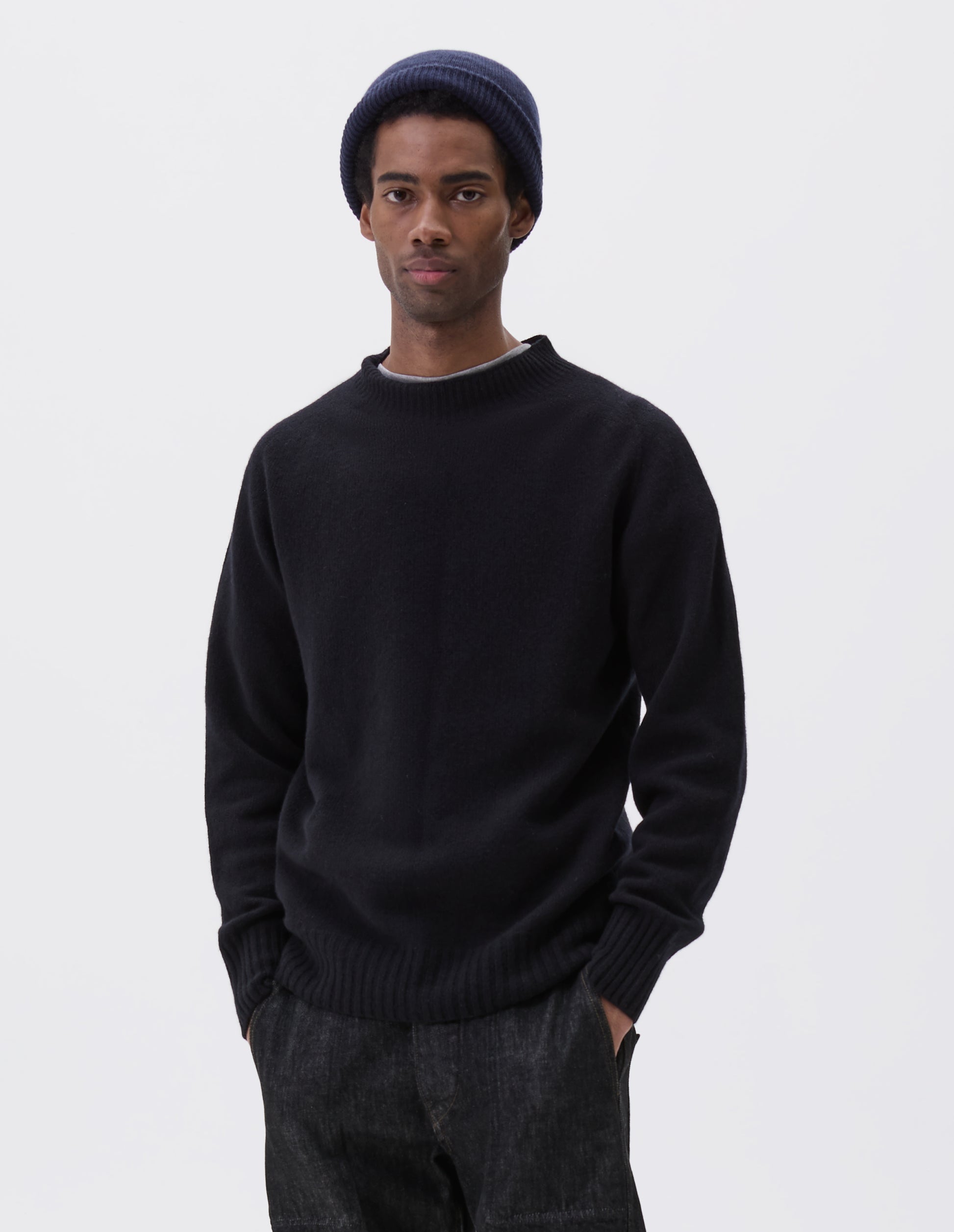 Cropped body image, front view of model wearing dark navy wool and cashmere jumper, with saddle sleeve, contrast rib on neck, cuffs and hem. Styled over grey t-shirt, with black denim jeans and navy blue beanie hat.