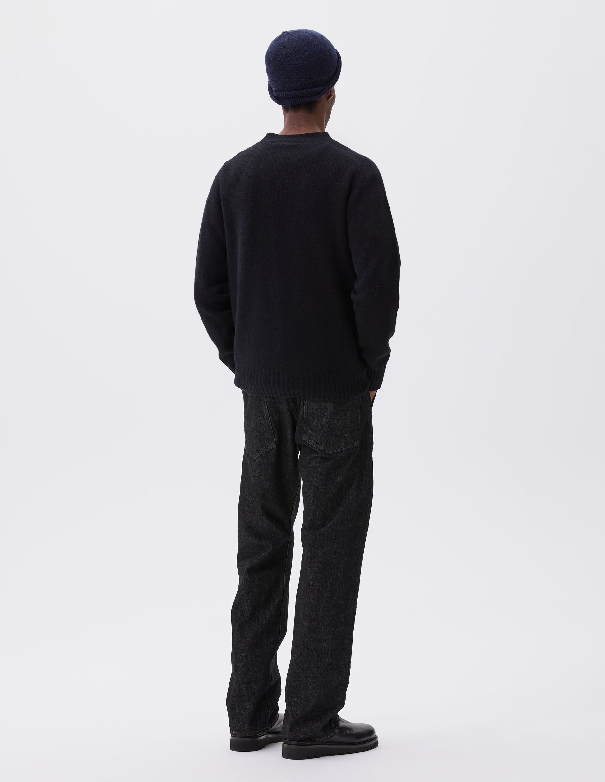 Full body image, back view of model wearing dark navy wool and cashmere jumper, with saddle sleeve, contrast rib on neck, cuffs and hem. Styled with navy blue beanie, black denim jeans and black leather boots.