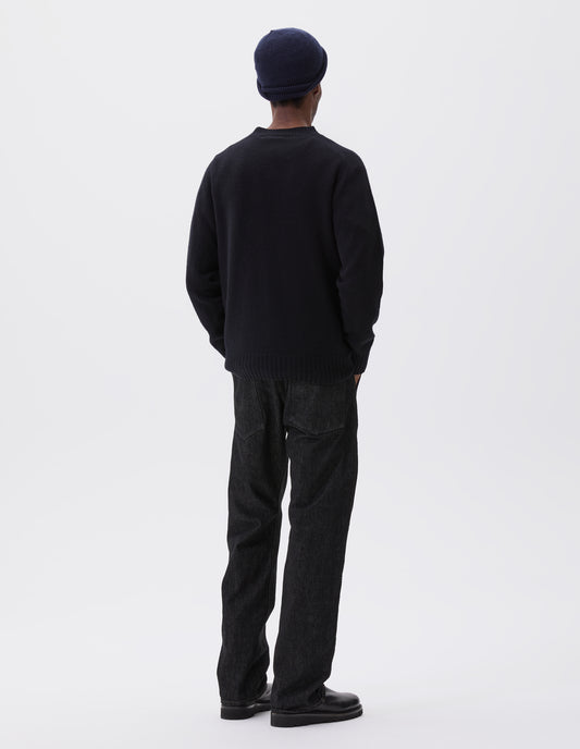 Full body image, back view of model wearing dark navy wool and cashmere jumper, with saddle sleeve, contrast rib on neck, cuffs and hem. Styled with navy blue beanie, black denim jeans and black leather boots.