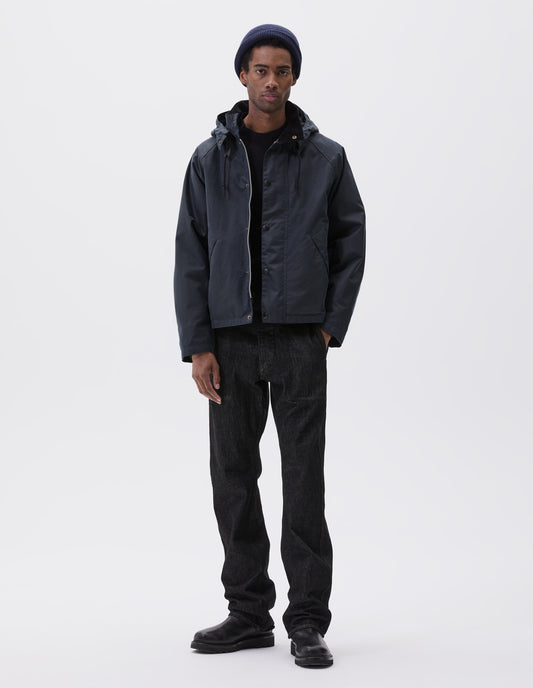 Full body image, front view of model wearing boxy dark blue jacket with corduroy collar and adjustable hood, raglan sleeve, two-way zip fastening, press stud storm flap and wide storm welt side pockets. Styled over black jumper with dark blue beanie, black jeans and black boots.