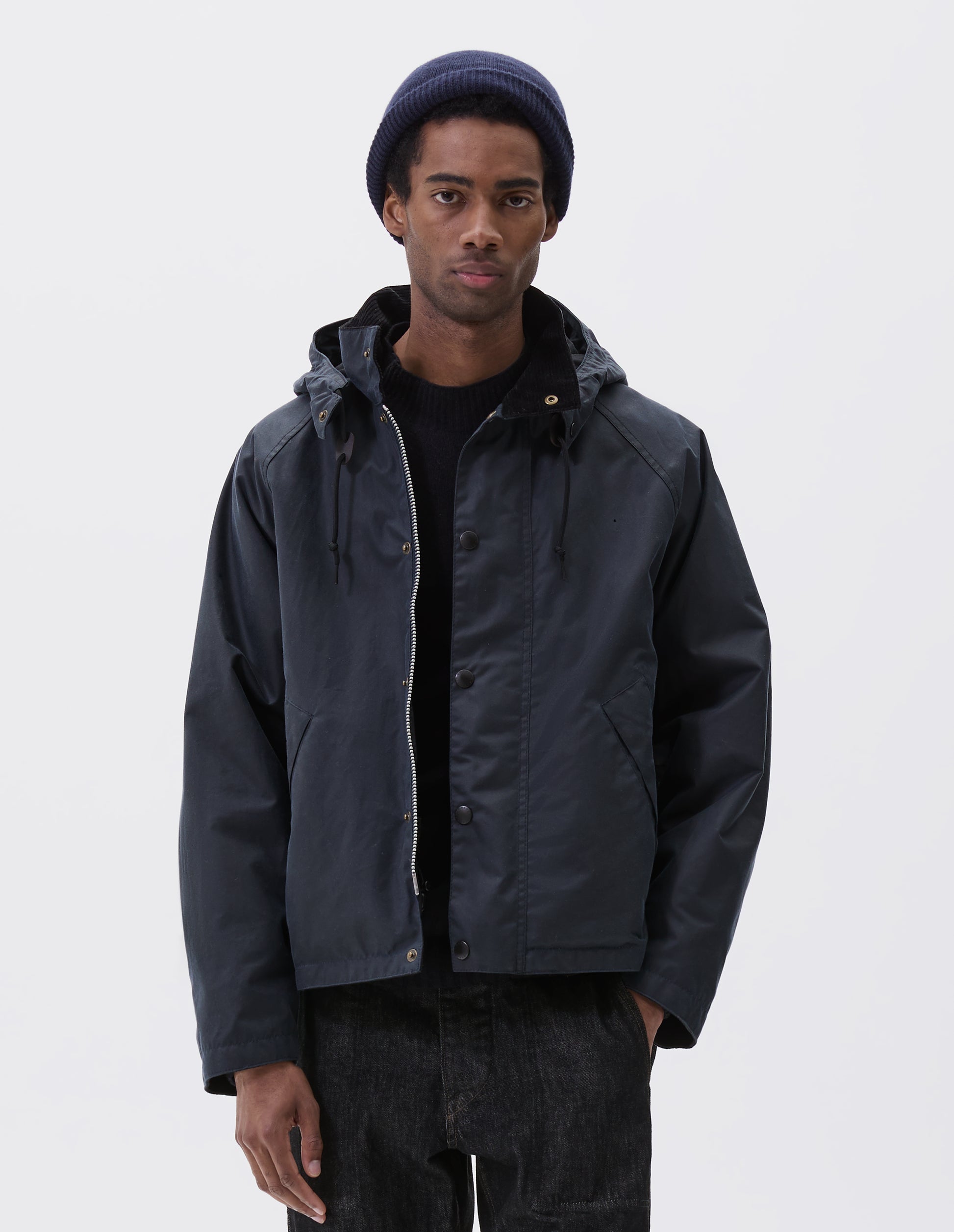 Cropped image, front view of model wearing boxy dark blue jacket with corduroy collar and adjustable hood, raglan sleeve, two-way zip fastening, press stud storm flap and wide storm welt side pockets. Styled over black jumper with dark blue beanie and black jeans.
