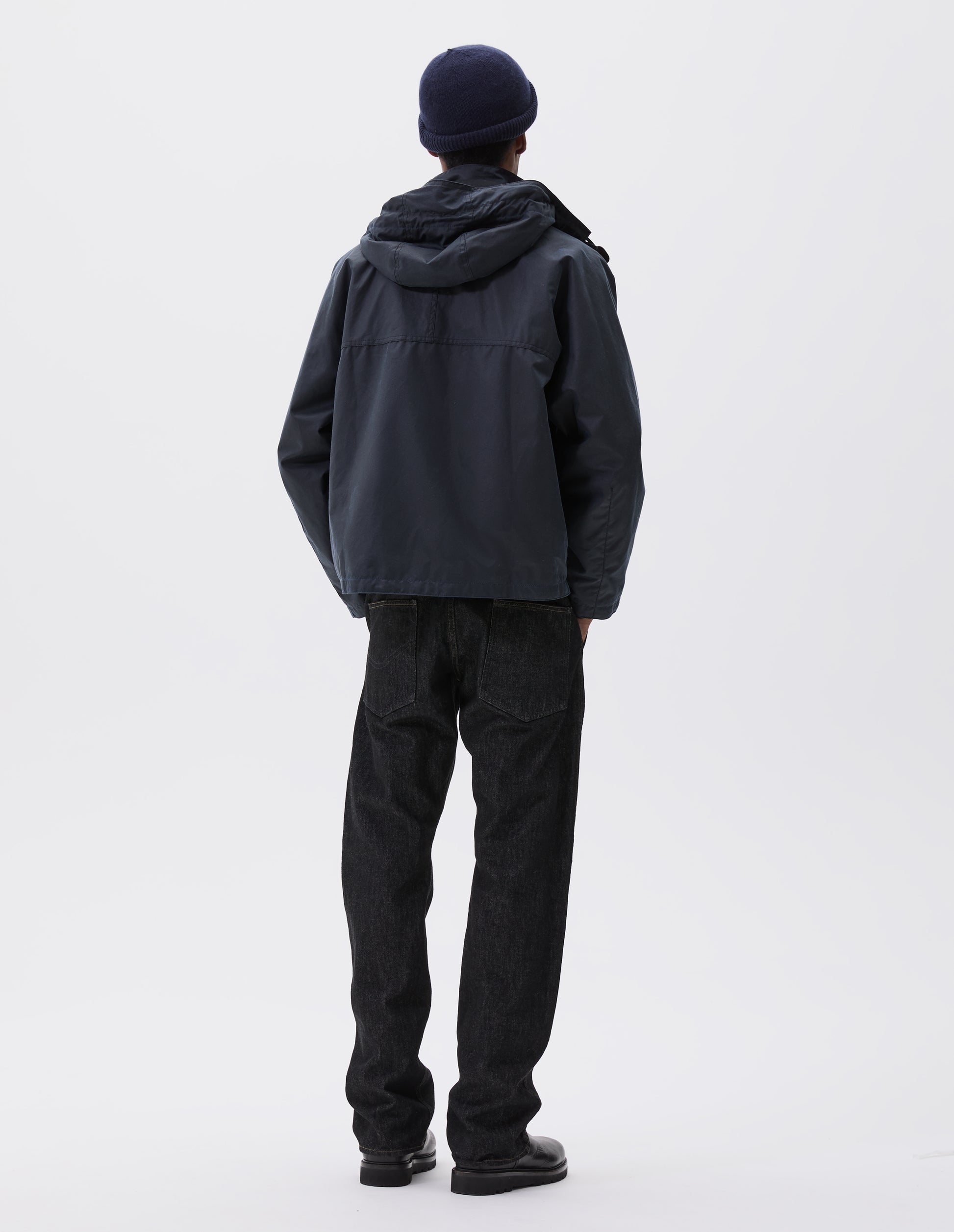 Full body image, back view of model wearing boxy dark blue jacket with raglan sleeve and hood with wire adjustable peak. Styled with dark blue beanie, black jeans and black boots.