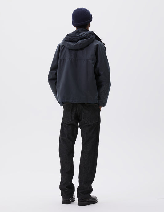 Full body image, back view of model wearing boxy dark blue jacket with raglan sleeve and hood with wire adjustable peak. Styled with dark blue beanie, black jeans and black boots.