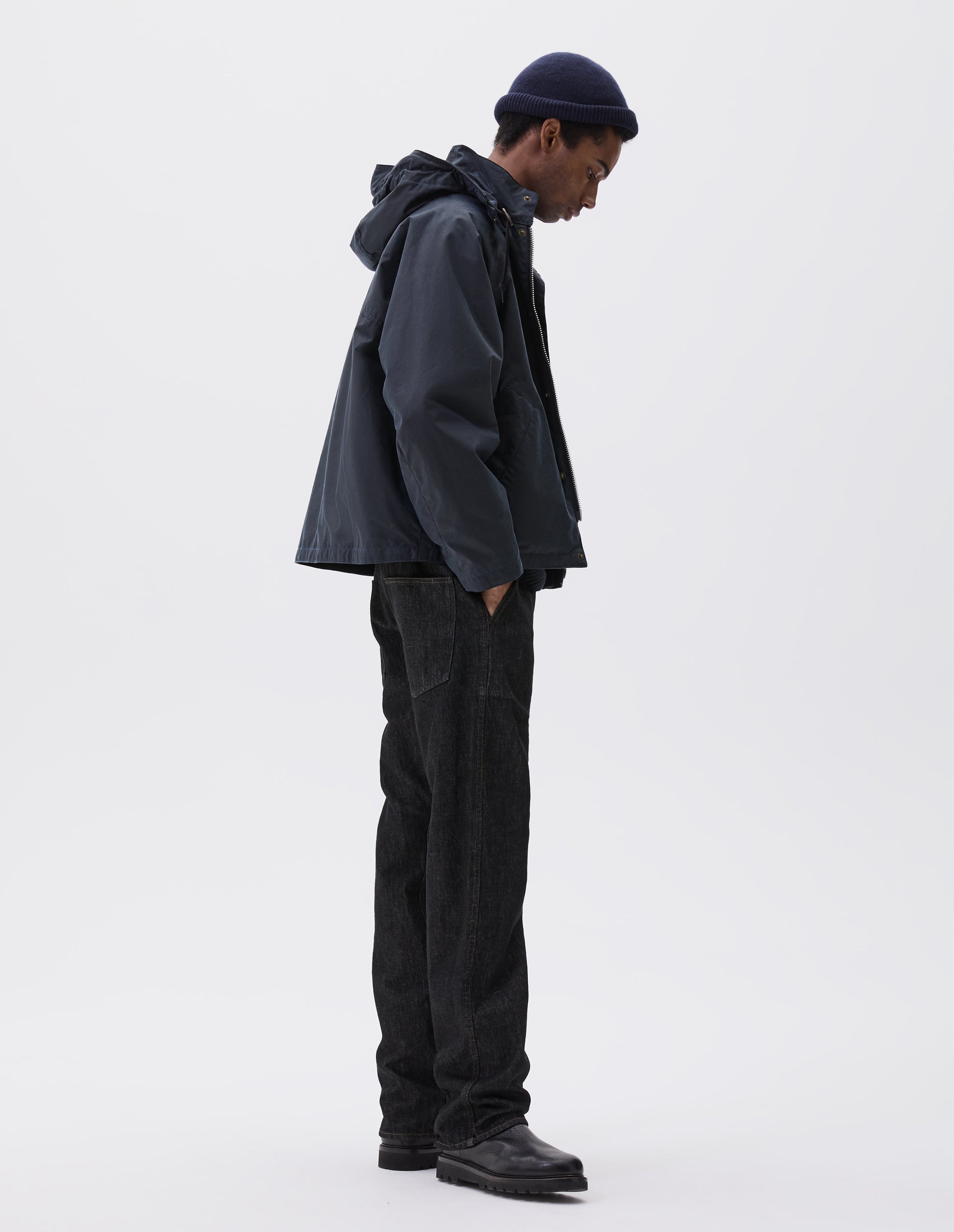 Full body image, side view of model wearing boxy dark blue jacket with stand collar and hood with wire adjustable peak and two way zip. Styled with dark blue beanie, black jeans and black boots.