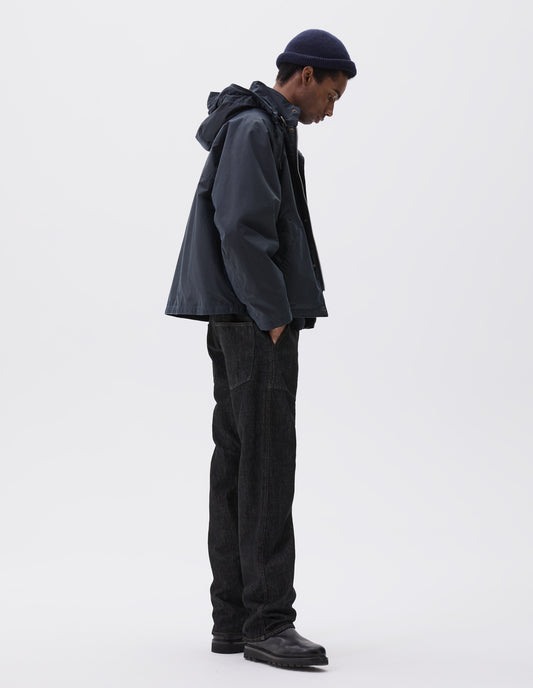 Full body image, side view of model wearing boxy dark blue jacket with stand collar and hood with wire adjustable peak and two way zip. Styled with dark blue beanie, black jeans and black boots.