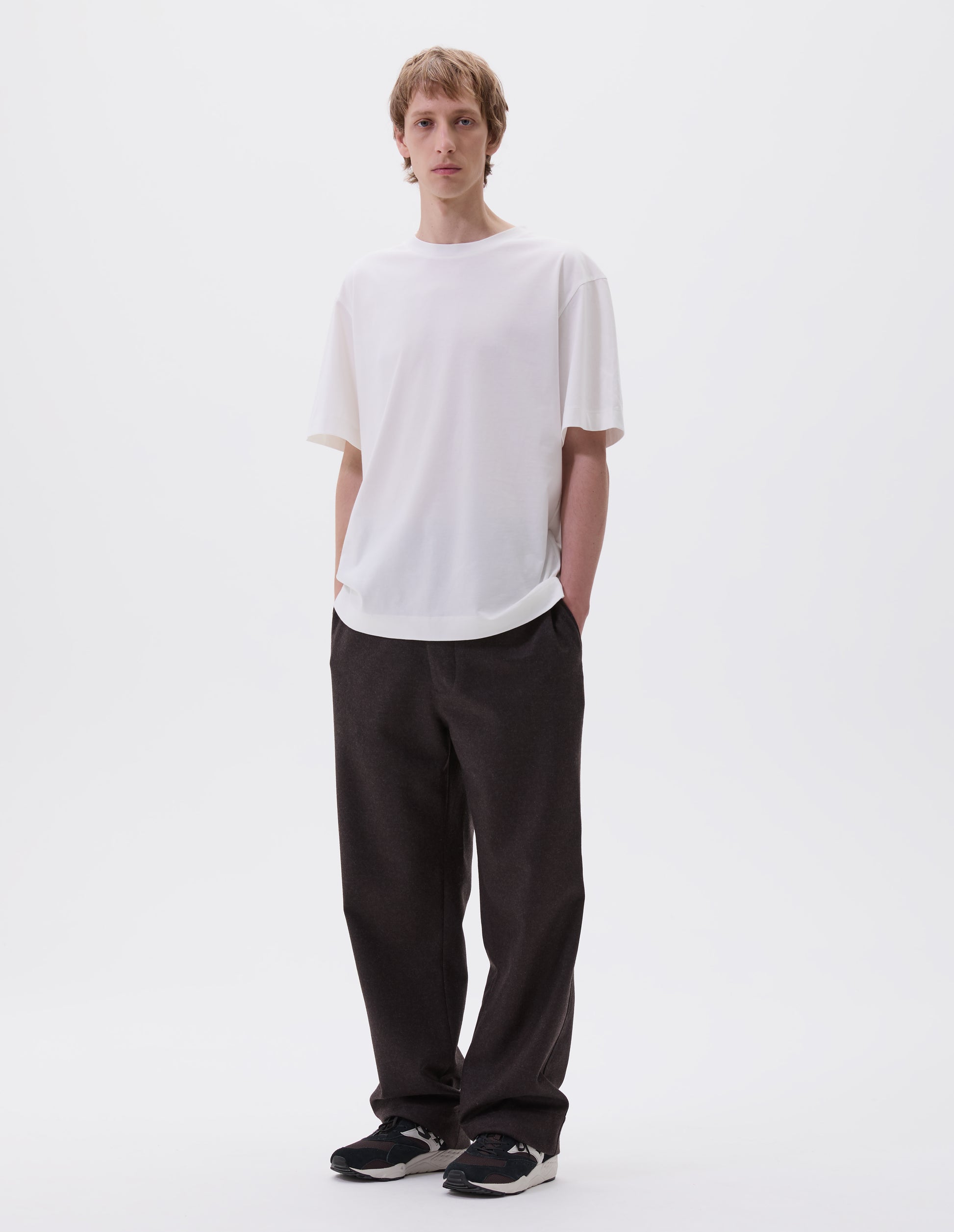 Full body image, front view of model wearing off white cotton short sleeve t-shirt. Styled with brown trousers and black and brown trainers.