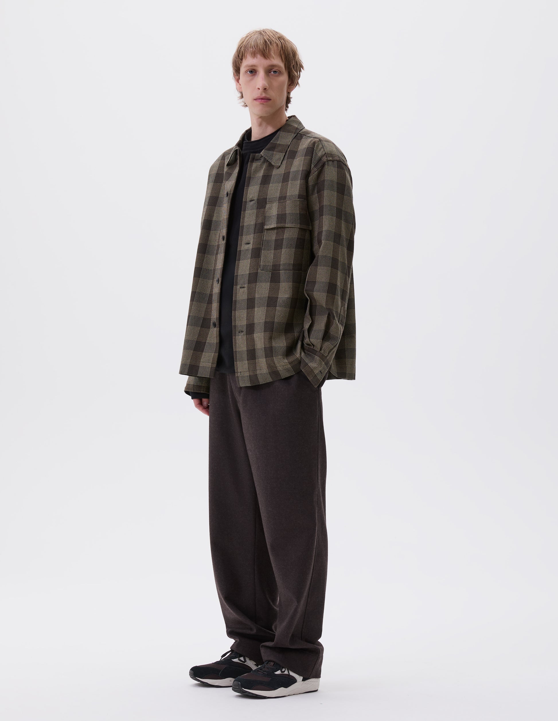 Full body image, front view of model wearing black, brown and green checked wool shirt with horn button fastening, deep placket and flap patch pocket. Styled over dark navy jumper, brown trousers and black and brown trainers.