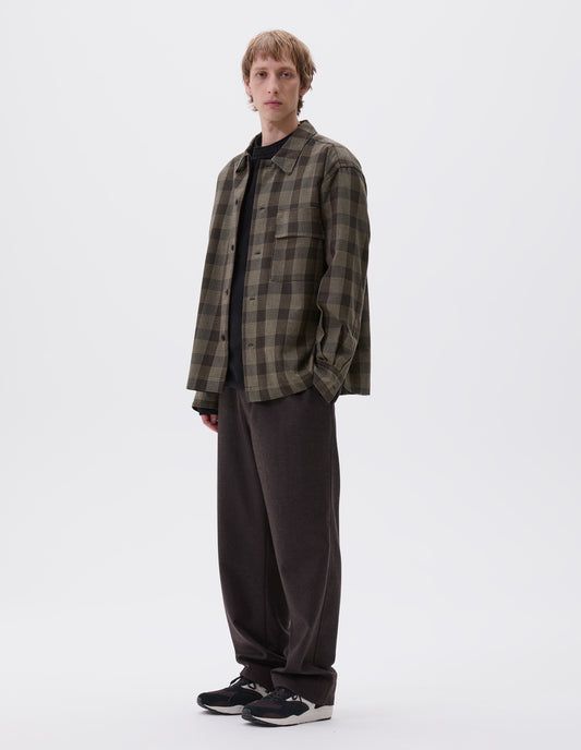 Full body image, front view of model wearing black, brown and green checked wool shirt with horn button fastening, deep placket and flap patch pocket. Styled over dark navy jumper, brown trousers and black and brown trainers.