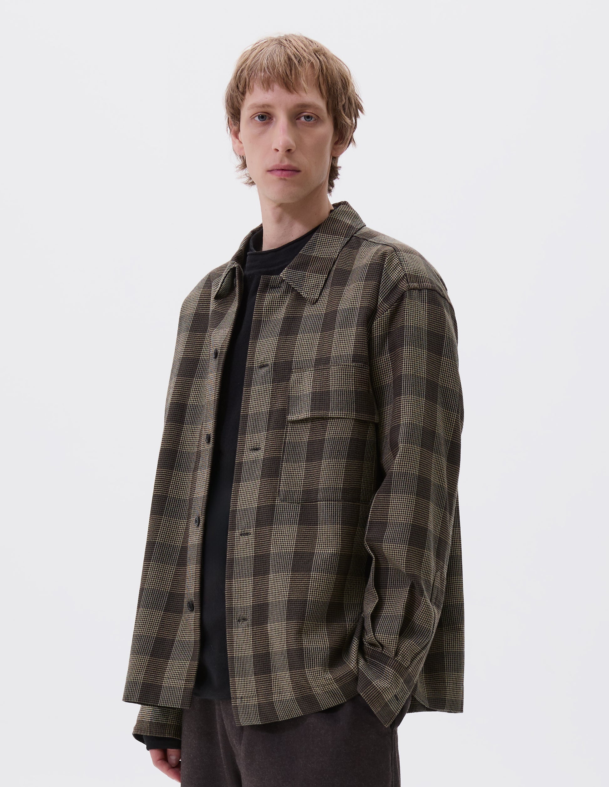 Cropped body image, front view of model wearing black, brown and green checked wool shirt with horn button fastening, deep placket and flap patch pocket. Styled over dark navy jumper and brown trousers.