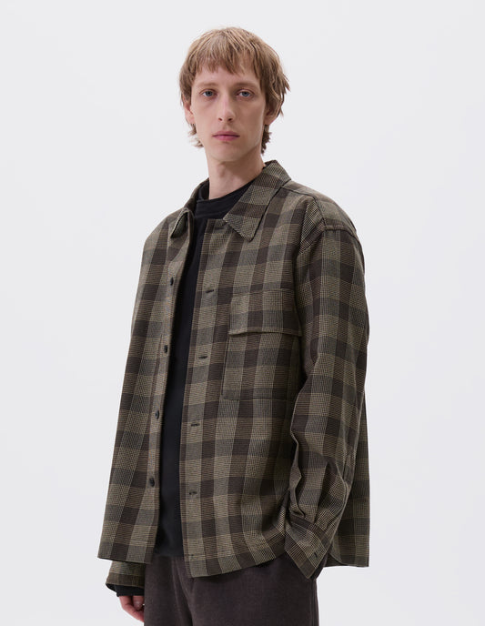 Cropped body image, front view of model wearing black, brown and green checked wool shirt with horn button fastening, deep placket and flap patch pocket. Styled over dark navy jumper and brown trousers.