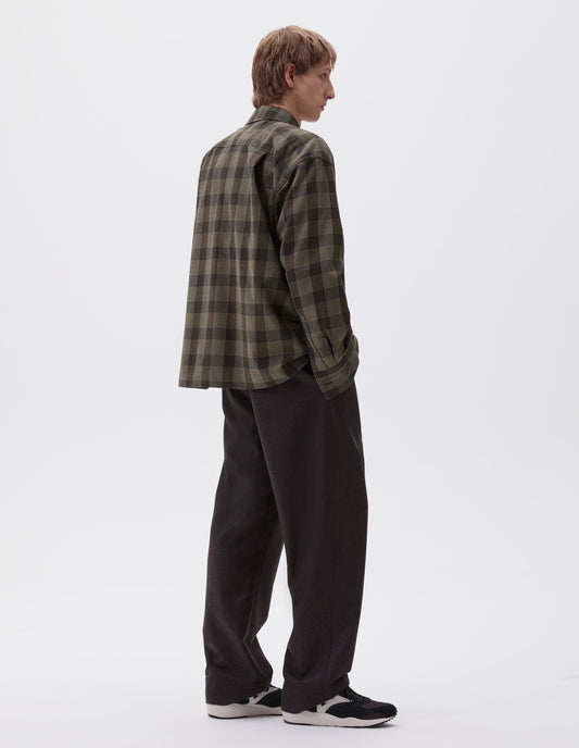 Full body image, back view of model wearing black, brown and green checked wool shirt, with horn button cuffs and box pleat on back yoke. Styled with brown trousers and black and brown trainers.