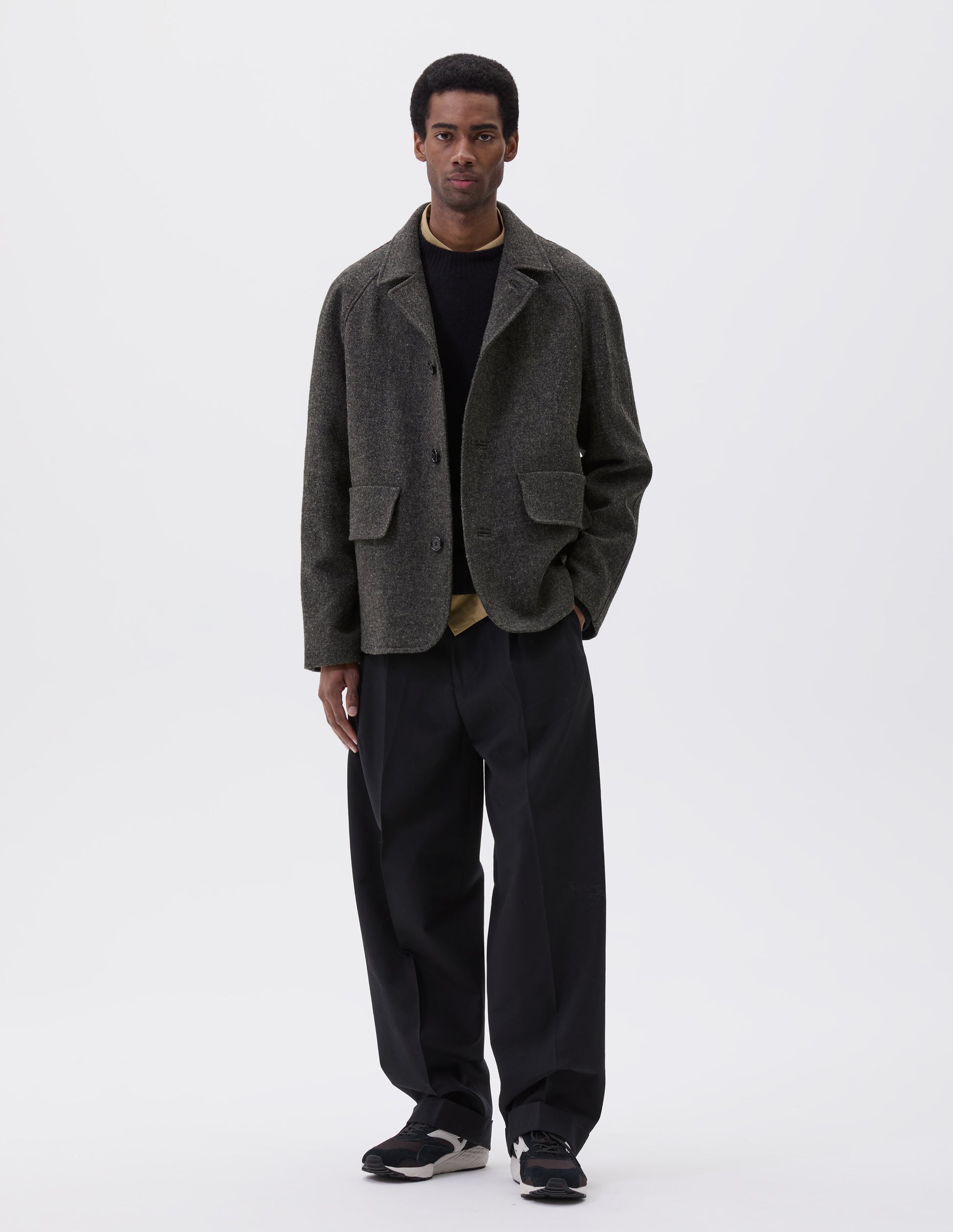 Full body image, front view of model wearing dark grey wool blazer with high collar fastening, deep flap welt pockets and subtle horn button detailing. Styled yellow shirt, black jumper, black trouser and brown and black trainers.