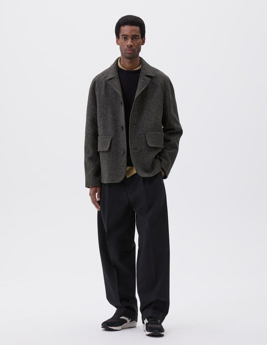 Full body image, front view of model wearing dark grey wool blazer with high collar fastening, deep flap welt pockets and subtle horn button detailing. Styled yellow shirt, black jumper, black trouser and brown and black trainers.