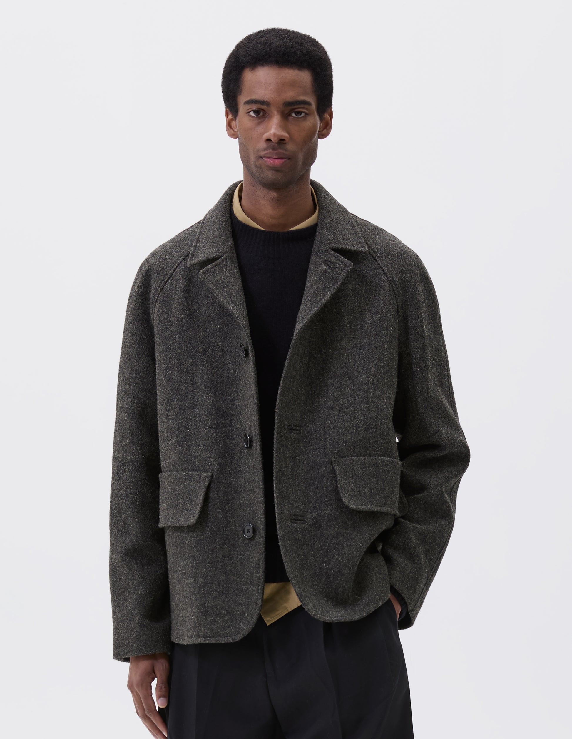 Cropped body image, front view of model wearing dark grey wool blazer with high collar fastening, deep flap welt pockets and subtle horn button detailing. Styled yellow shirt, black jumper and black trouser.