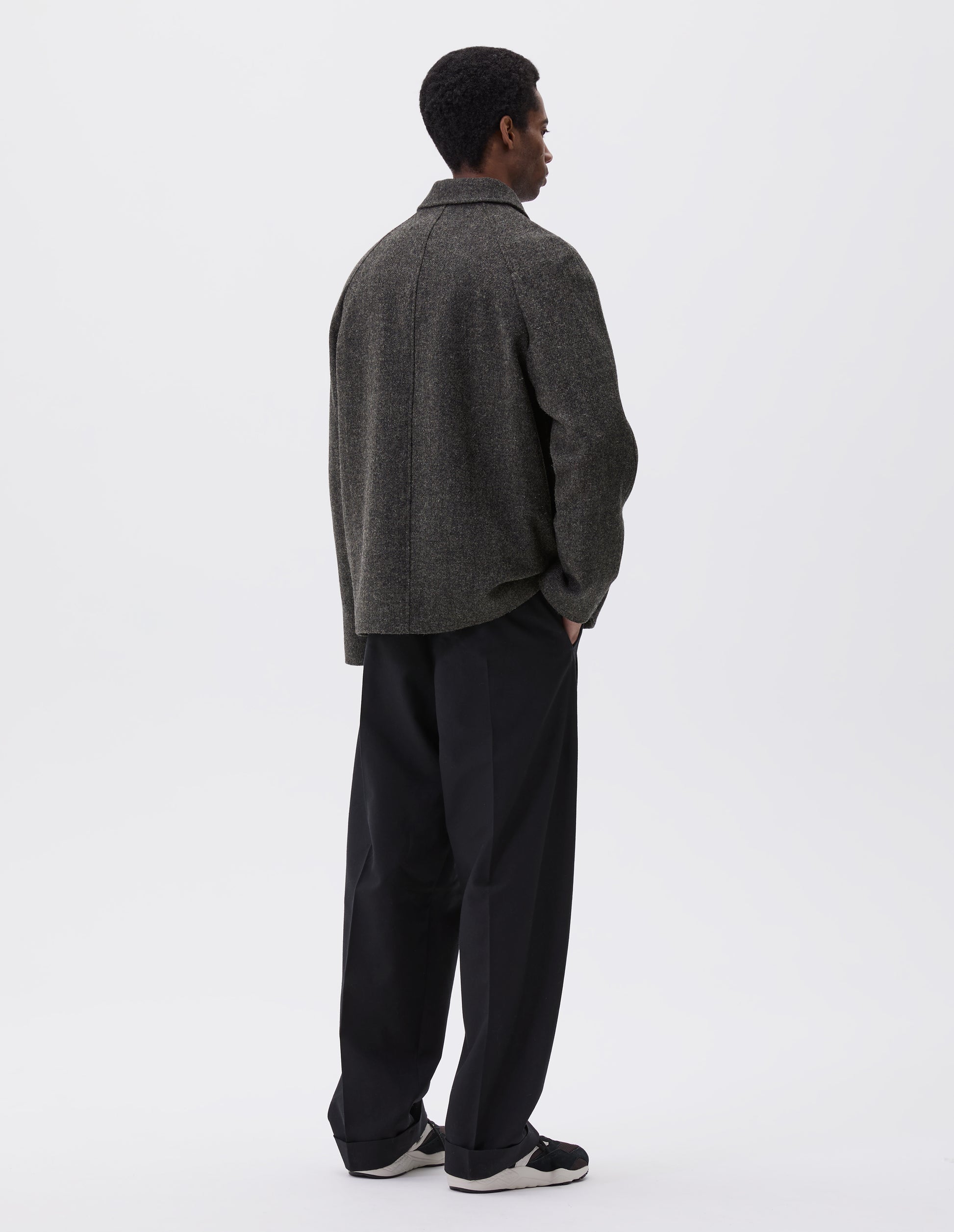 Full body image, back view of model wearing dark grey wool blazer with high collar fastening and raglan sleeve Styled with black trousers and brown and black trainers.