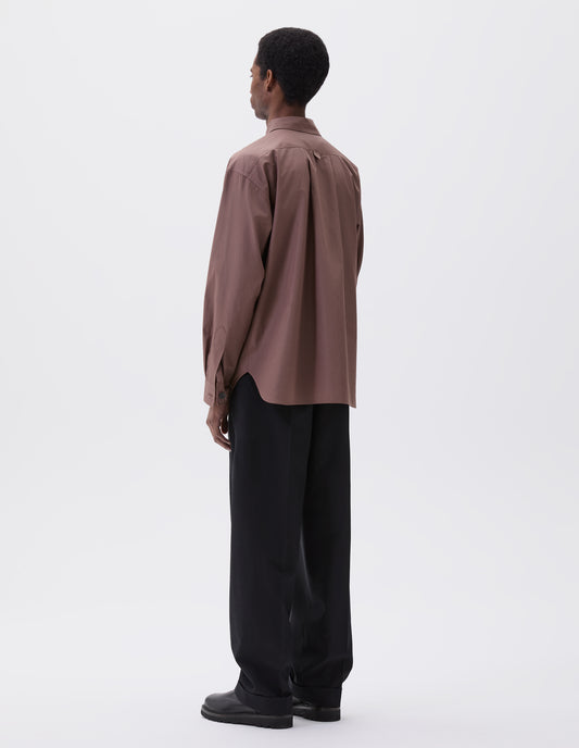 Full body image, back view of model wearing relaxed dark pink shirt with hanger loop detail on back, split yoke and mother of pearl buttons on cuffs. Styled with black trousers and black leather boots.