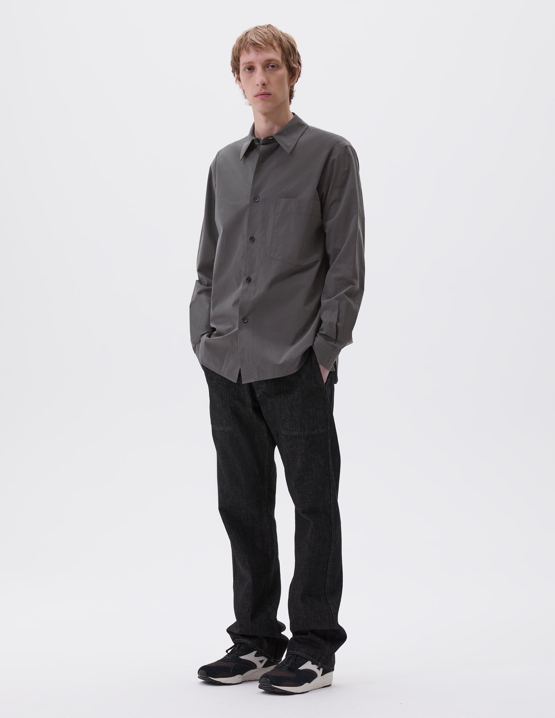 Full body image, front view of model wearing regular cut dark grey shirt with chest pocket and horn buttons. Styled with black jeans and black, brown and white trainers.