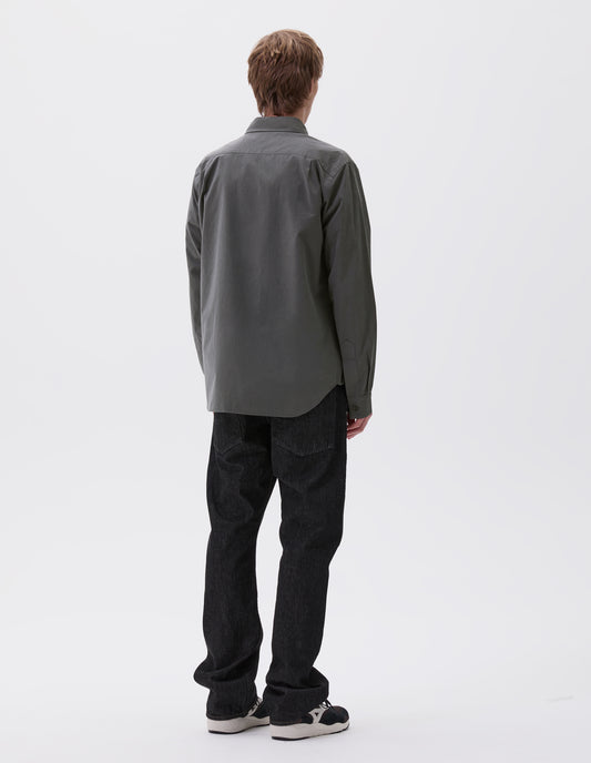 Full body image, back view of model wearing regular cut dark grey shirt with curved hem, pleats on back yoke and horn buttons on cuffs. Styled with black jeans and black, brown and white trainers.