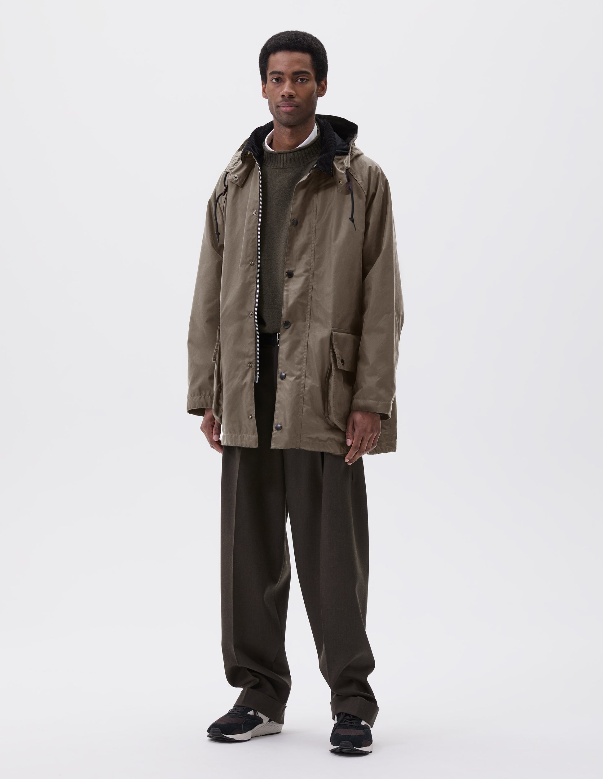 Full body image, front view of model wearing oversized dark beige parka with corduroy collar and hood with wire adjustable peak, raglan sleeve, two way zip fastening, press stud fastening storm flap and large bellows pockets with press stud fastening envelope closure. Styled over green jumper with white shirt, black leather belt, brown trousers and black, brown and white trainers. 