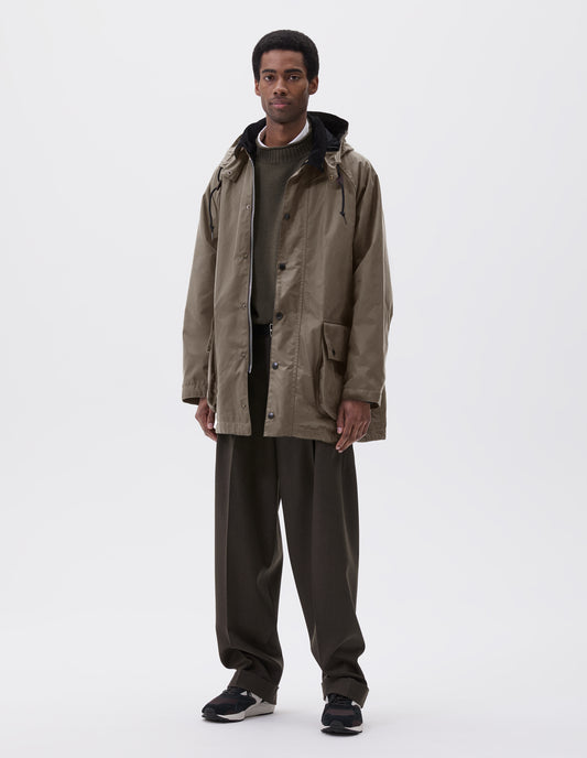 Full body image, front view of model wearing oversized dark beige parka with corduroy collar and hood with wire adjustable peak, raglan sleeve, two way zip fastening, press stud fastening storm flap and large bellows pockets with press stud fastening envelope closure. Styled over green jumper with white shirt, black leather belt, brown trousers and black, brown and white trainers. 