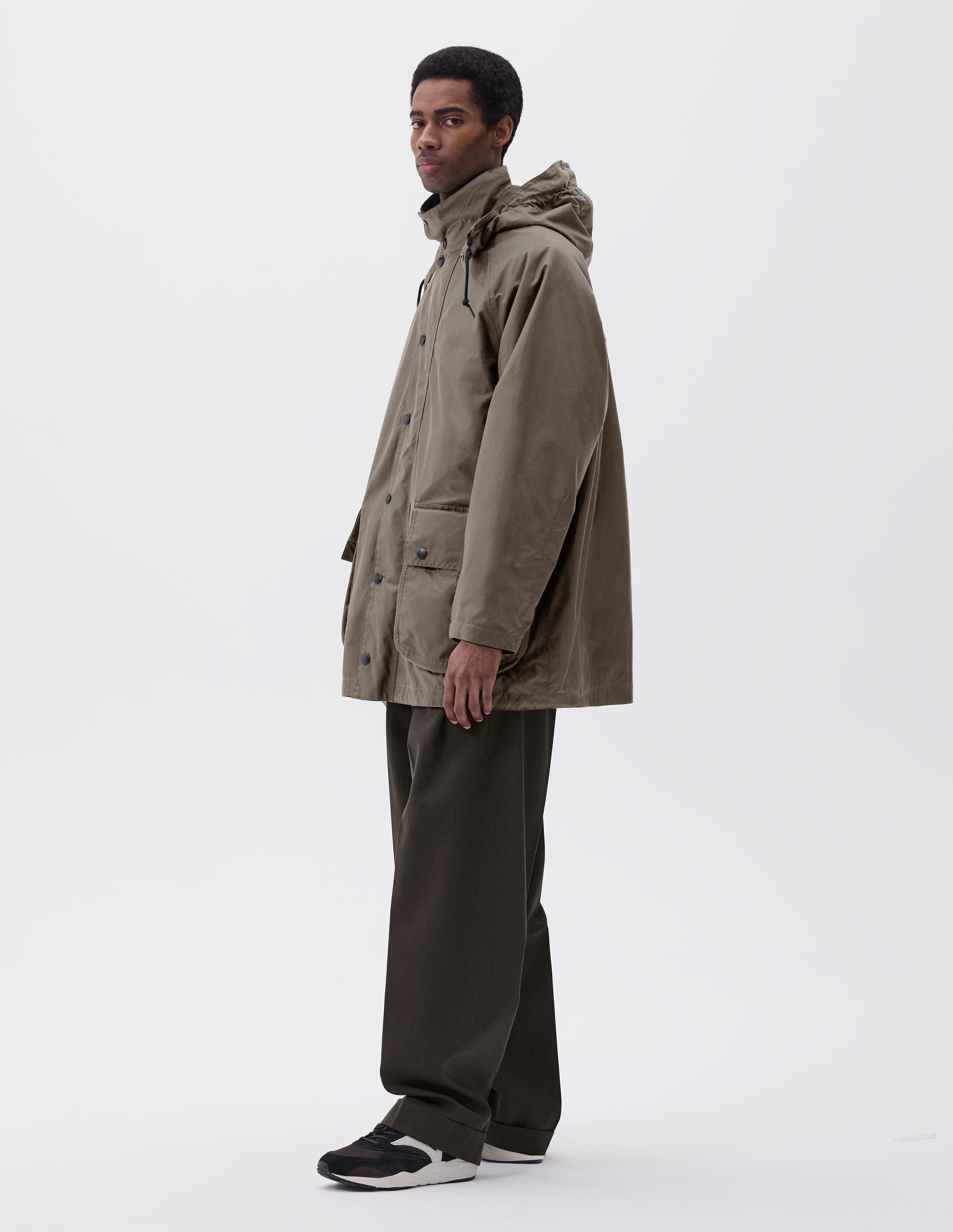 Full body image, side view of model wearing oversized dark beige parka with stand collar and hood with wire adjustable peak, raglan sleeve, press stud fastening storm flap and large bellows pockets with press stud fastening envelope closure. Styled with brown trousers and black, brown and white trainers. 