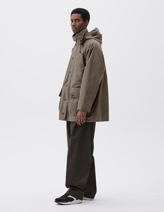 Full body image, side view of model wearing oversized dark beige parka with stand collar and hood with wire adjustable peak, raglan sleeve, press stud fastening storm flap and large bellows pockets with press stud fastening envelope closure. Styled with brown trousers and black, brown and white trainers. 