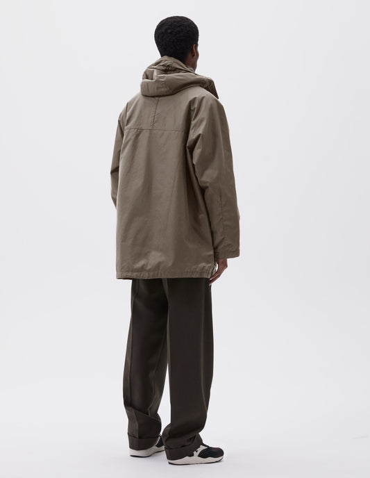 Full body image, back view of model wearing oversized dark beige parka with hood and raglan sleeves. Styled with brown trousers and black, brown and white trainers. 