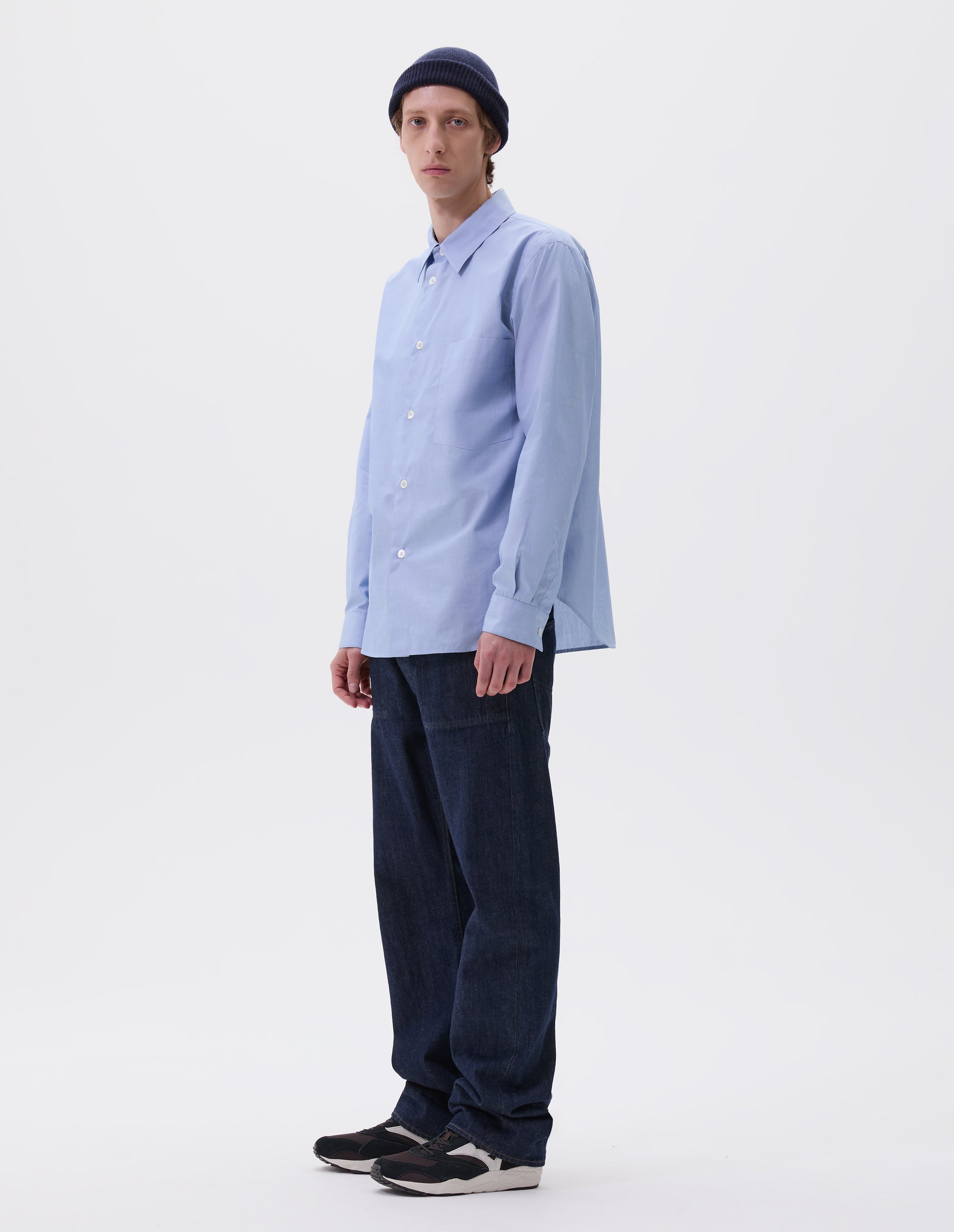 Full body image, front view of model wearing pale blue cotton shirt with chest pocket, curved hem and mother of pearl buttons. Styled with navy blue beanie hat, indigo blue denim jeans and black and brown trainers.