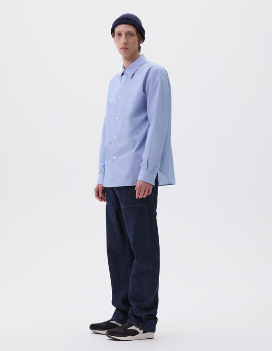 Full body image, front view of model wearing pale blue cotton shirt with chest pocket, curved hem and mother of pearl buttons. Styled with navy blue beanie hat, indigo blue denim jeans and black and brown trainers.