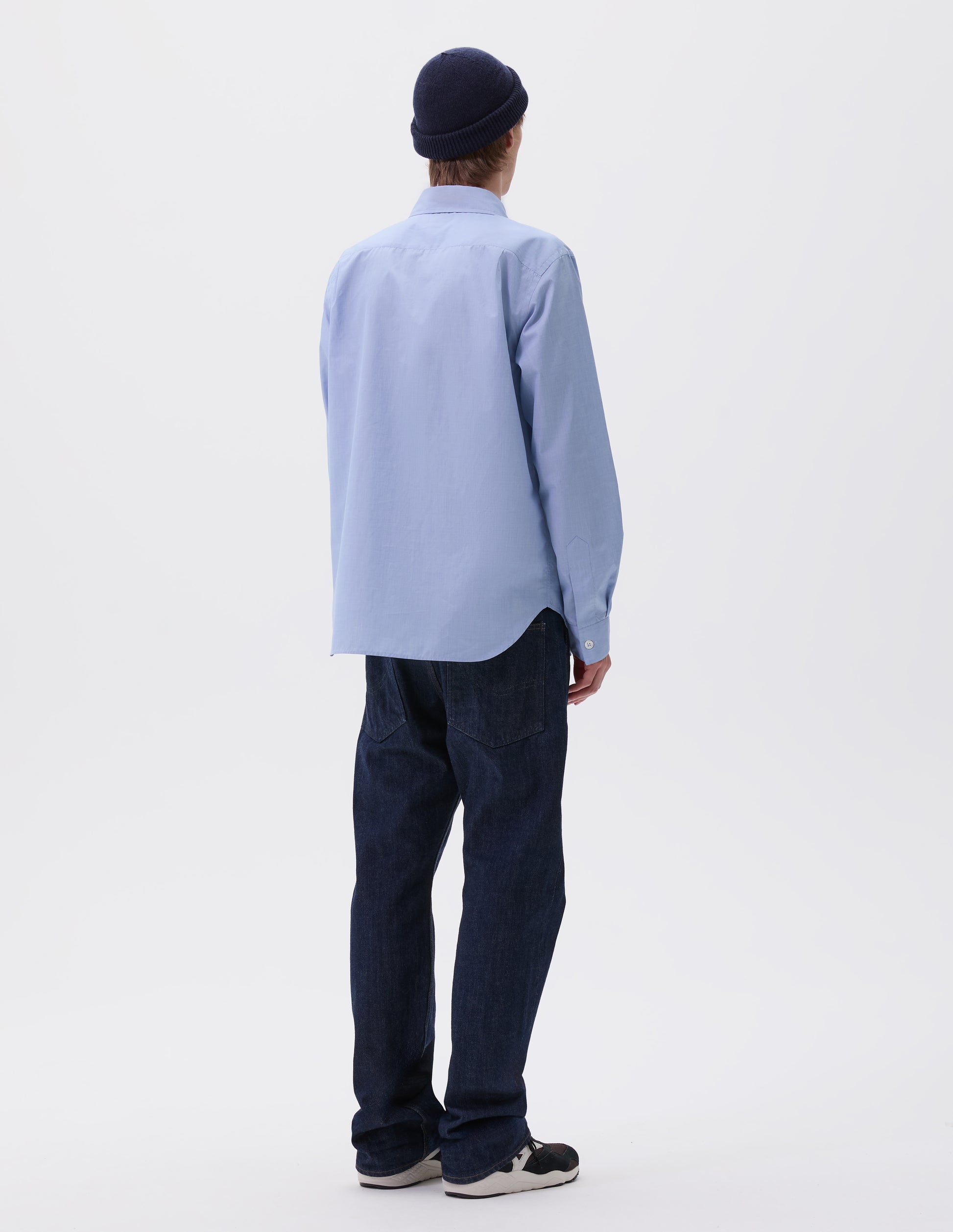 Full body image, back view of model wearing pale blue cotton shirt, with curved hem, pleats on back yoke and mother of pearl button cuffs. Styled with navy blue beanie hat, indigo blue denim jeans and black and brown trainers.