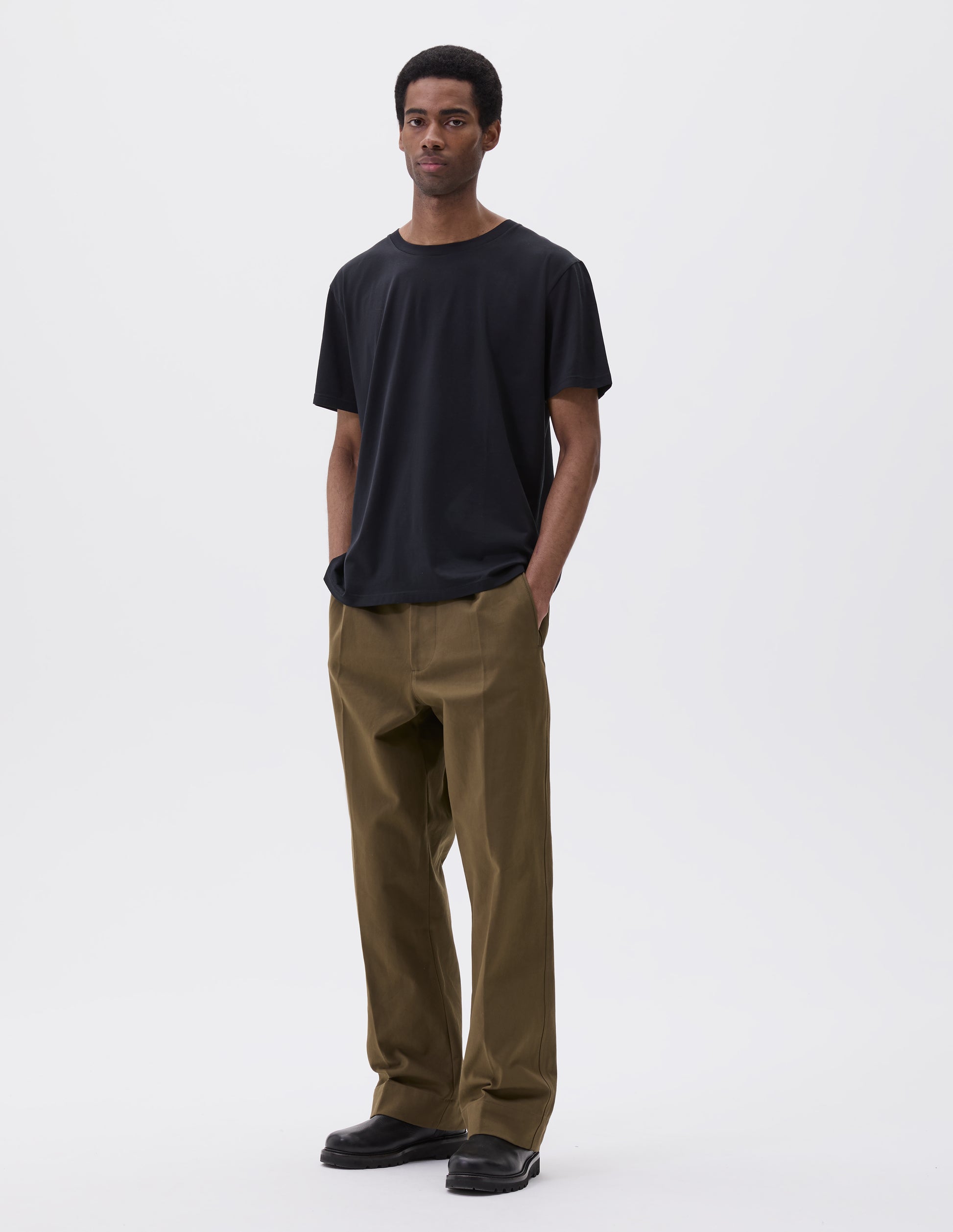 Full body image, front view of model wearing black cotton short sleeve t-shirt. Styled with green trousers and black leather boots.
