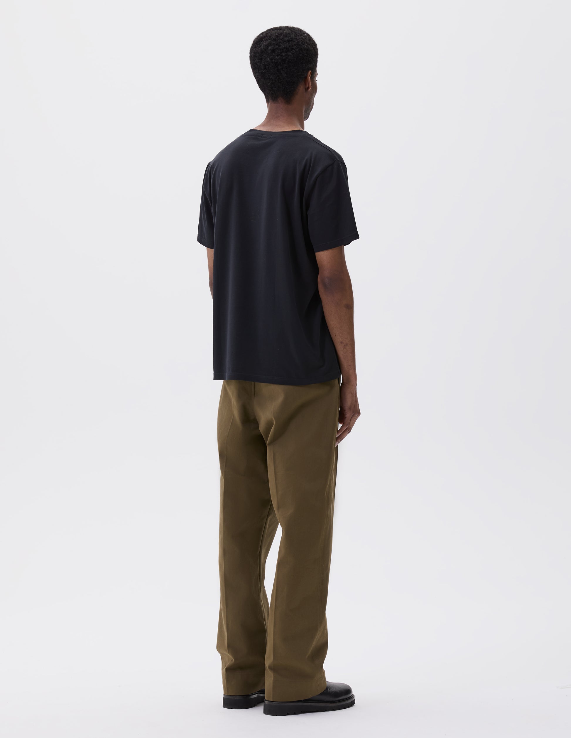 Full body image, back view of model wearing black cotton short sleeve t-shirt. Styled with green trousers and black leather boots.