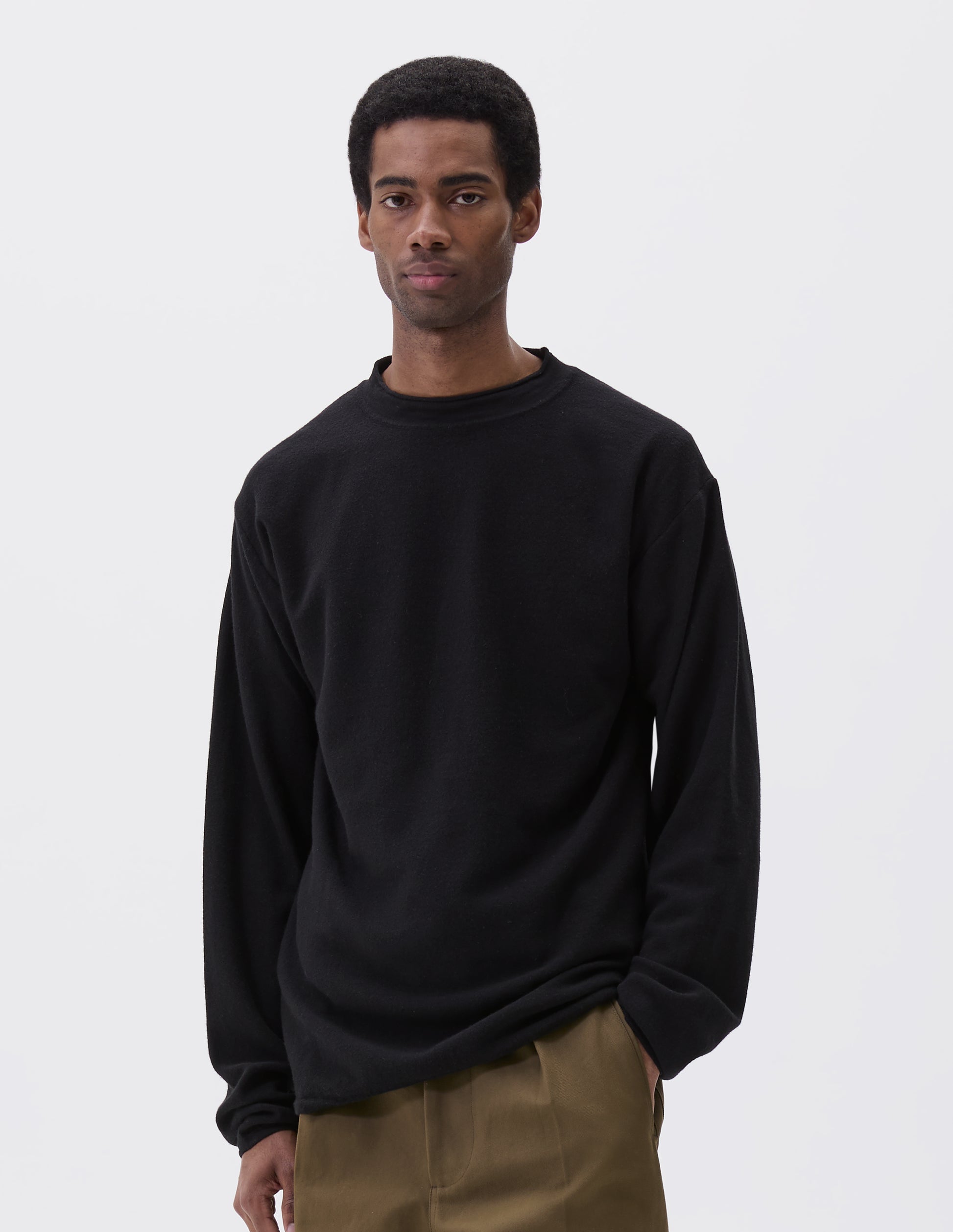 Cropped image, front view of model wearing relaxed black crew neck jumper with rolled edge detailing on neck, rib trim on cuffs and hem. Styled with khaki green trousers.