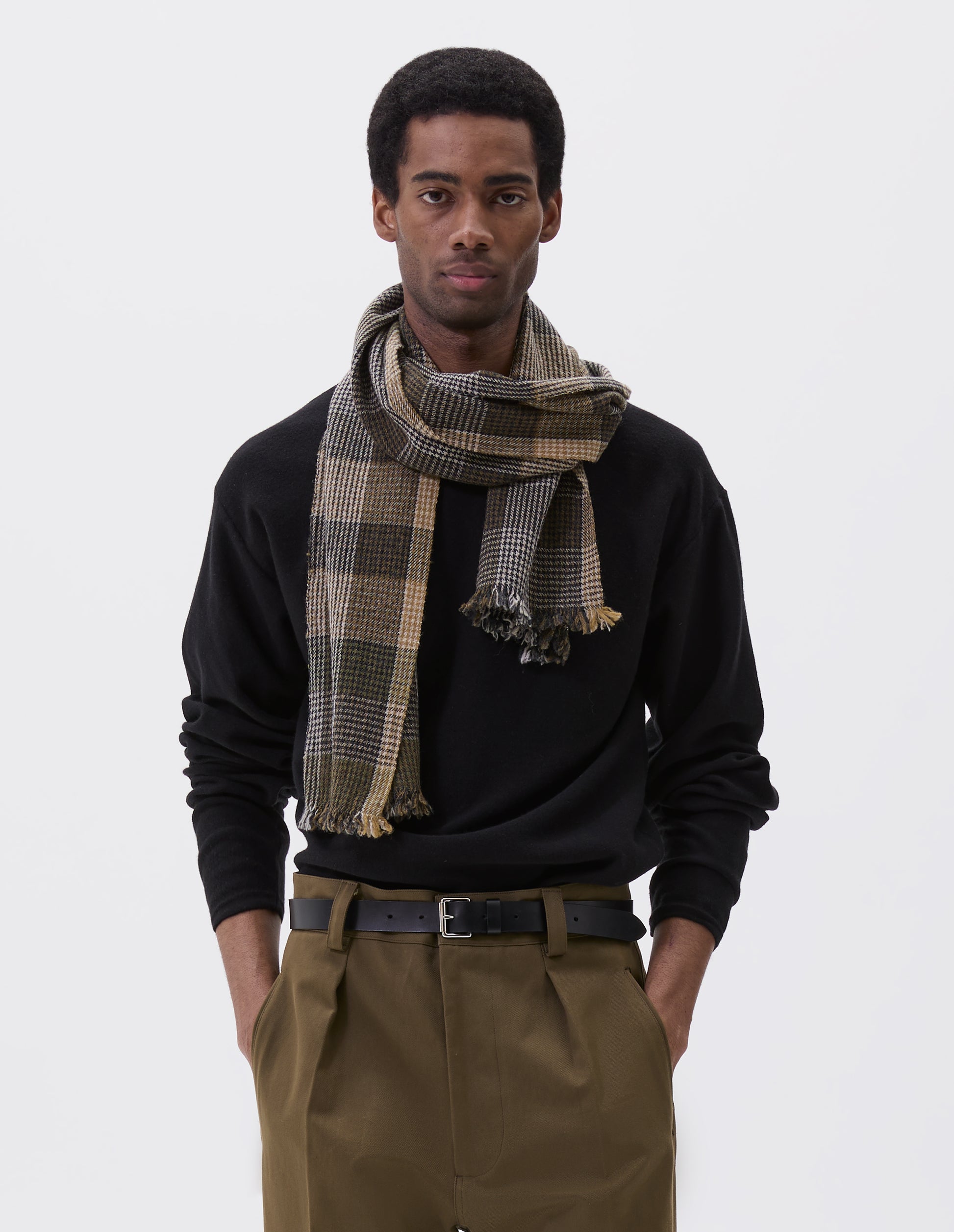 Cropped body image, front view of model wearing handwoven puppytooth overcheck scarf in khaki and black with hints of white and yellow. Styled with black long sleeve jumper, black leather belt and green pleated trousers.