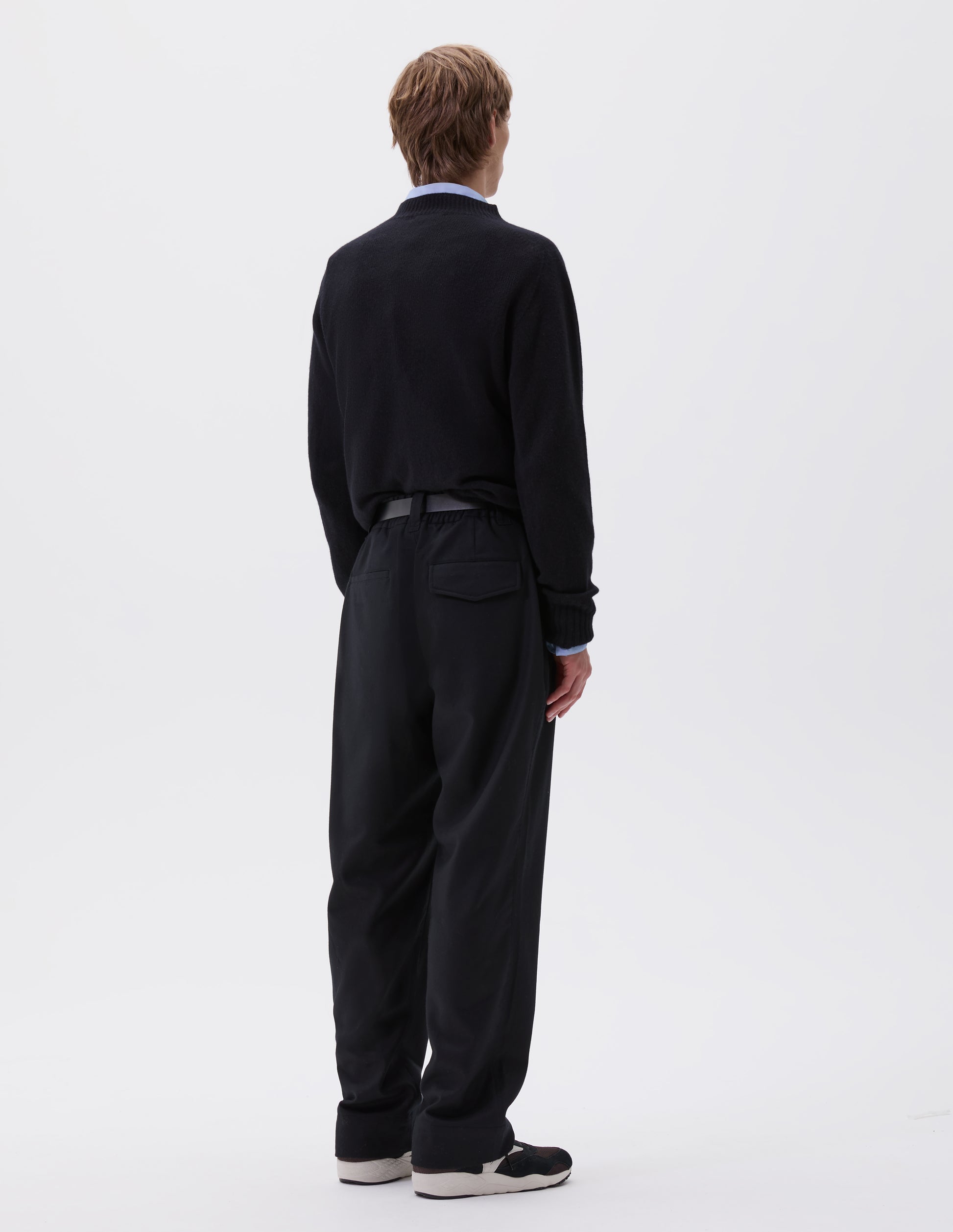 Full body image, back view of model wearing navy blue trousers, with half elasticated waistband, belt loops and welt and concealed button fastening flap back pockets. Styled with blue shirt, navy jumper, black leather belt and black and brown trainers.