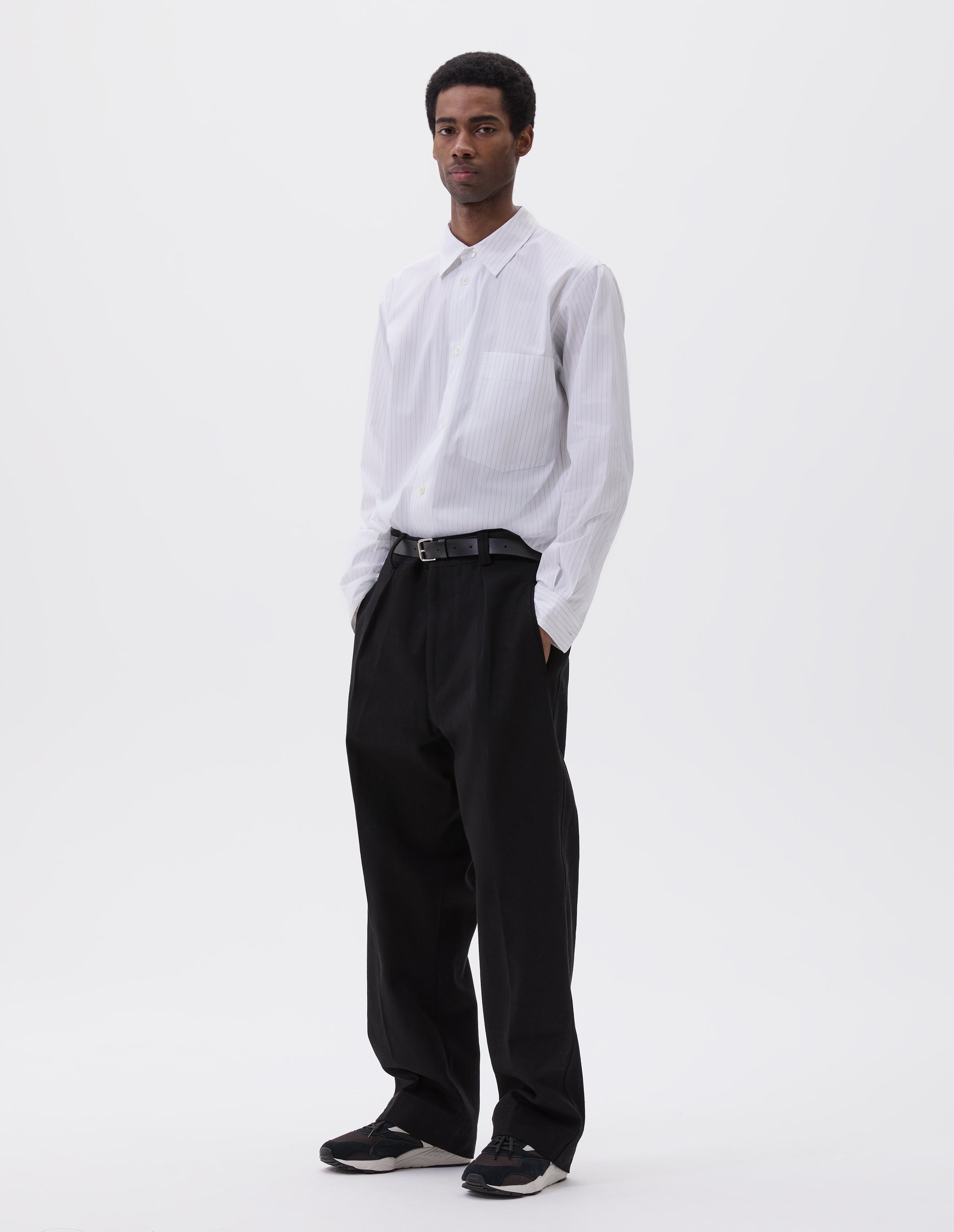 Full body image, front view of model wearing regular cut white shirt with grey stripes, chest pocket and mother of pearl buttons. Styled with black trousers, black leather belt and black, brown and white trainers.