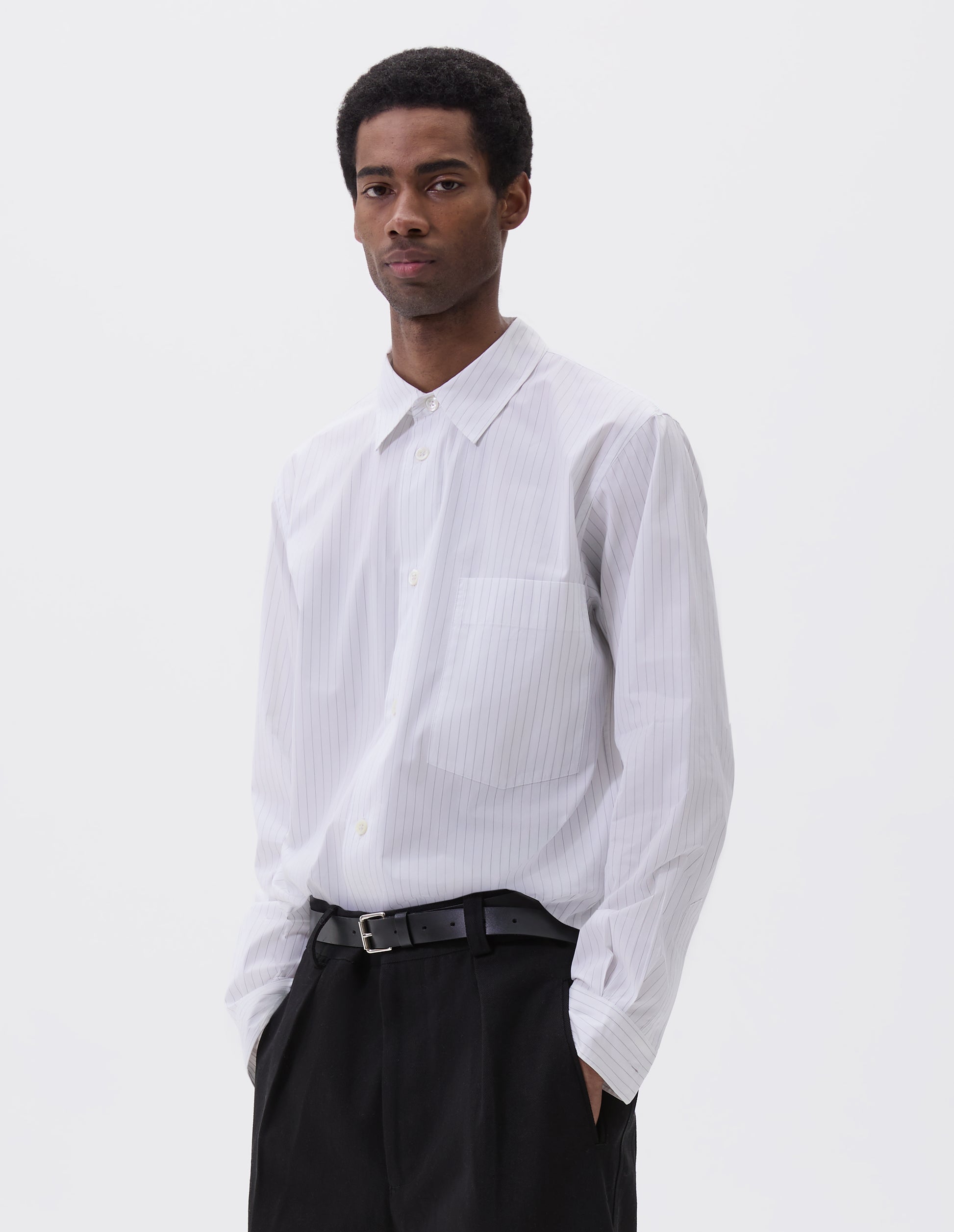Cropped image, front view of model wearing regular cut white shirt with grey stripes, chest pocket and mother of pearl buttons. Styled with black trousers and black leather belt.