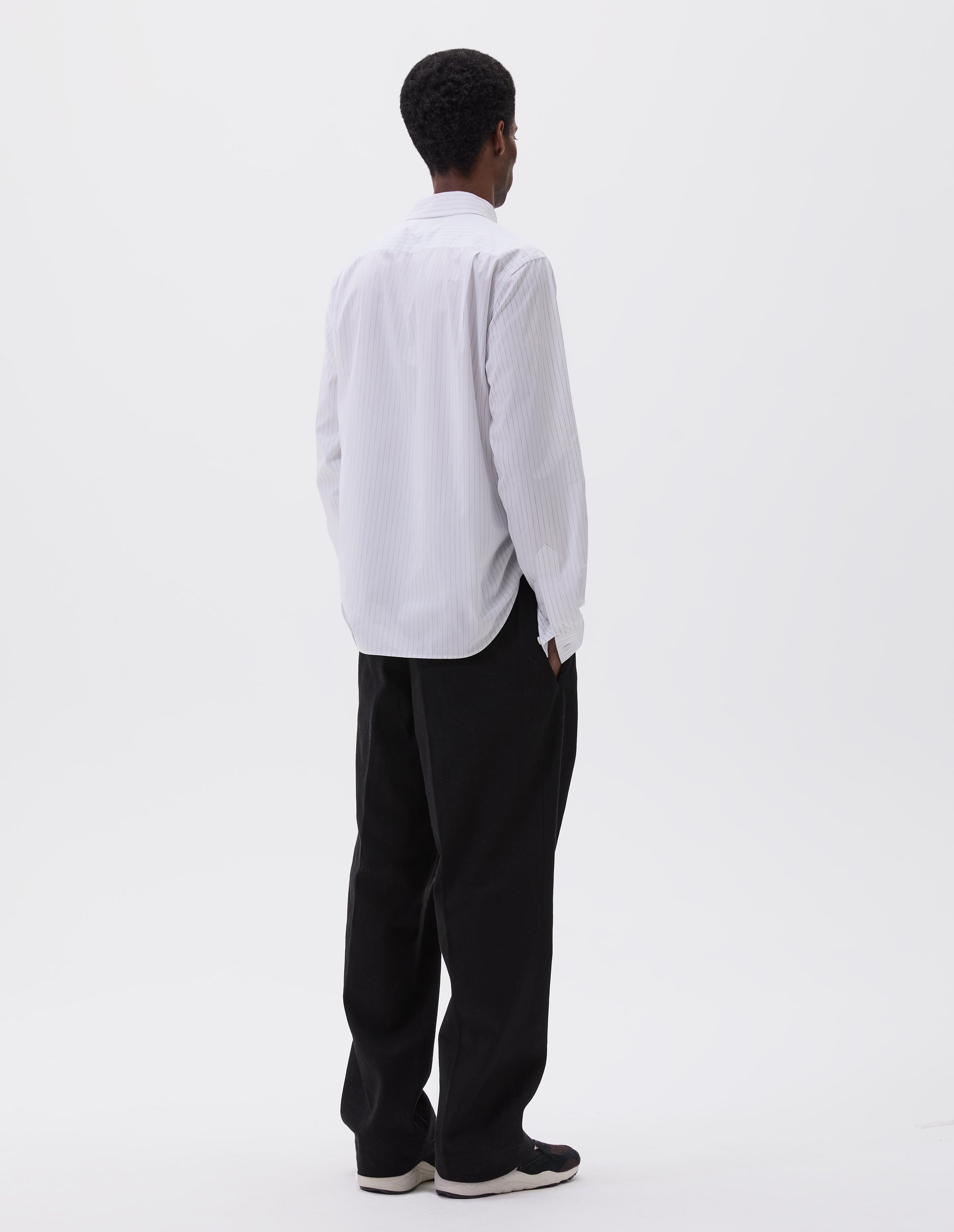 Full body image, back view of model wearing black plainweave wool trousers with concealed side pockets and deep button through welt back pocket. Styled with white and grey striped cotton shirt and black and brown trainers.