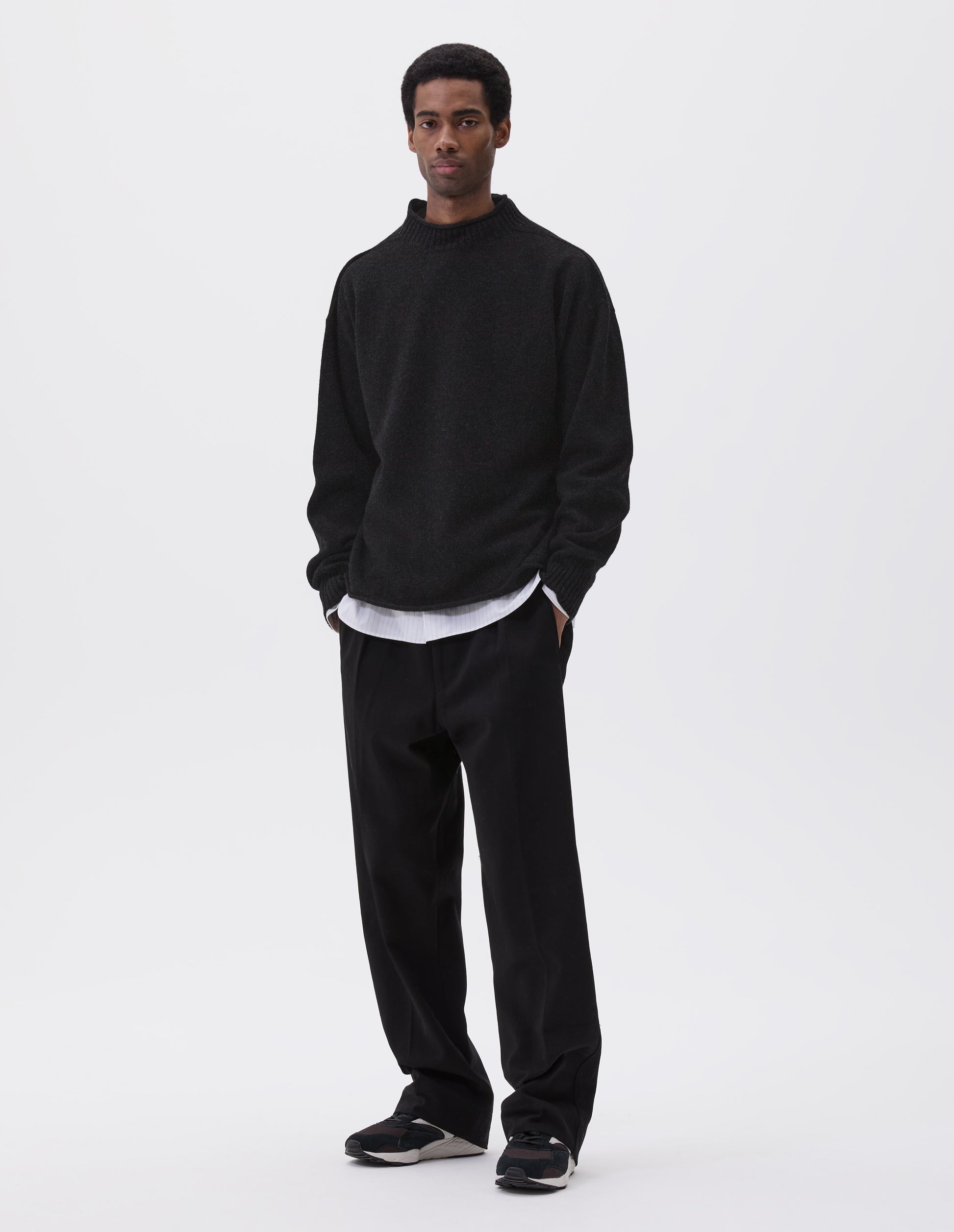 Full body image, front view of model wearing black long sleeve jumper with rolled edge neck and hem and contrasting rib on neck and sleeves. Styled with black trousers, white and grey striped shirt and black and brown trainers.