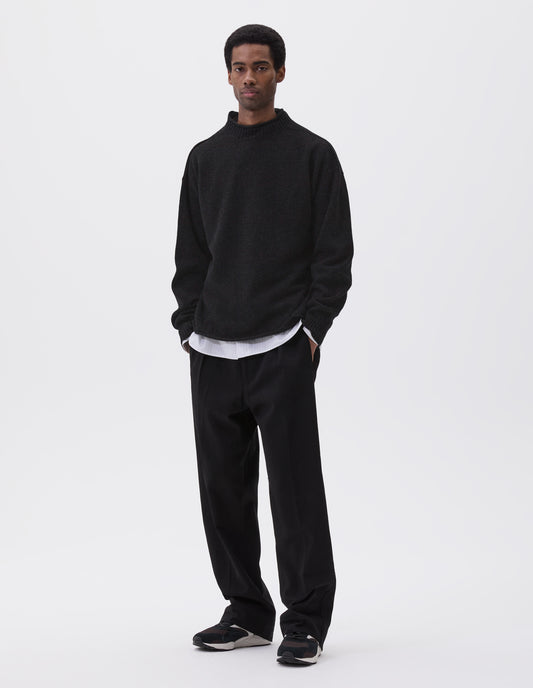 Full body image, front view of model wearing black long sleeve jumper with rolled edge neck and hem and contrasting rib on neck and sleeves. Styled with black trousers, white and grey striped shirt and black and brown trainers.