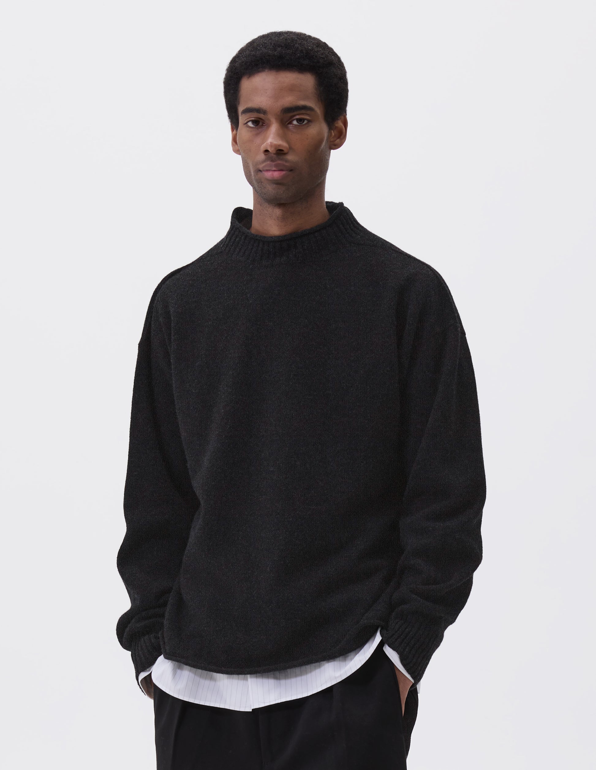 Cropped body image, front view of model wearing black long sleeve jumper with rolled edge neck and hem and contrasting rib on neck and sleeves. Styled with black trousers and white and grey striped shirt.
